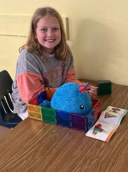 Girl smiling, holding a stuffed narwhal in a toy construction.