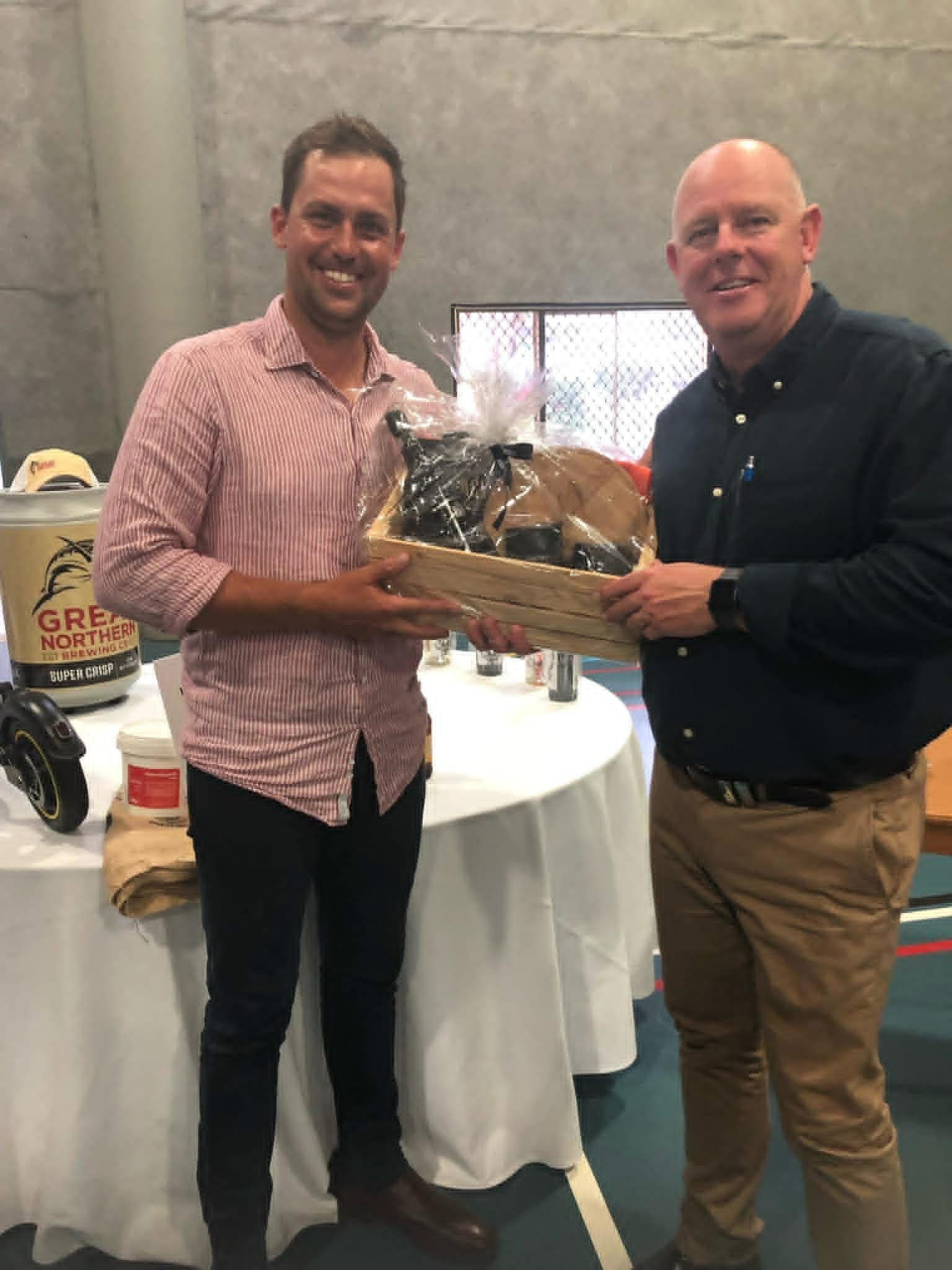 Two men smiling and presenting a gift basket at a table