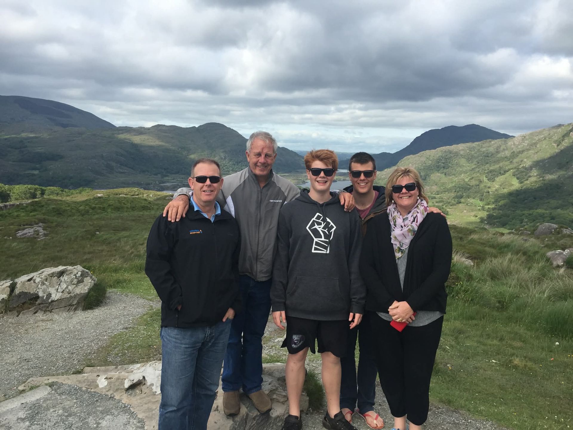 Five people posing on a mountain overlook with green hills and cloudy sky behind them