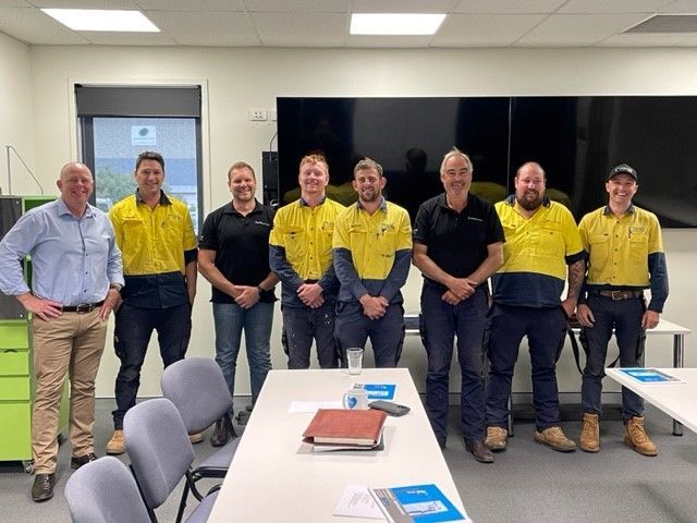 Seven people posing in a conference room, four in yellow shirts and three in dark shirts, standing behind tables and chairs.