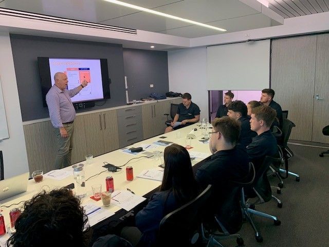 Team meeting in a conference room, with a presenter pointing at a screen while others watch around a table