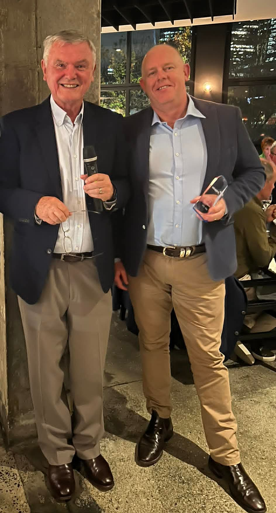 Two men in blazers holding drinks and awards at an indoor evening event.