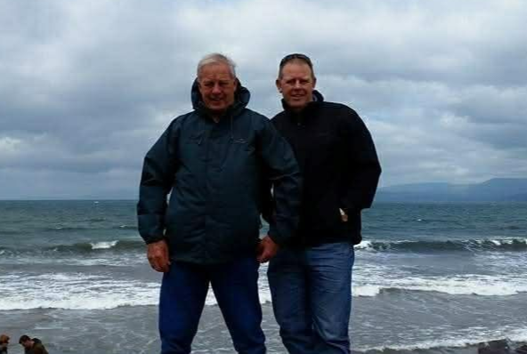 Two people standing on a windy beach with ocean waves and cloudy sky behind them