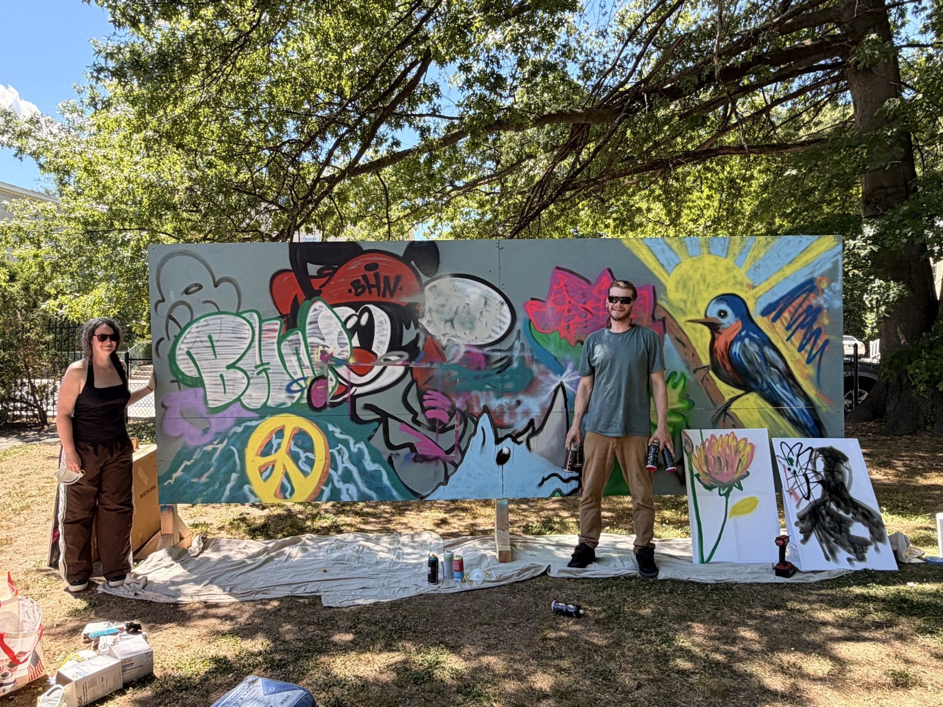 Two adults stand in front of the completed community graffiti board. The finished board was decorated with smiling characters, peace signs, a rhino, a blue bird, positive phrases, like 