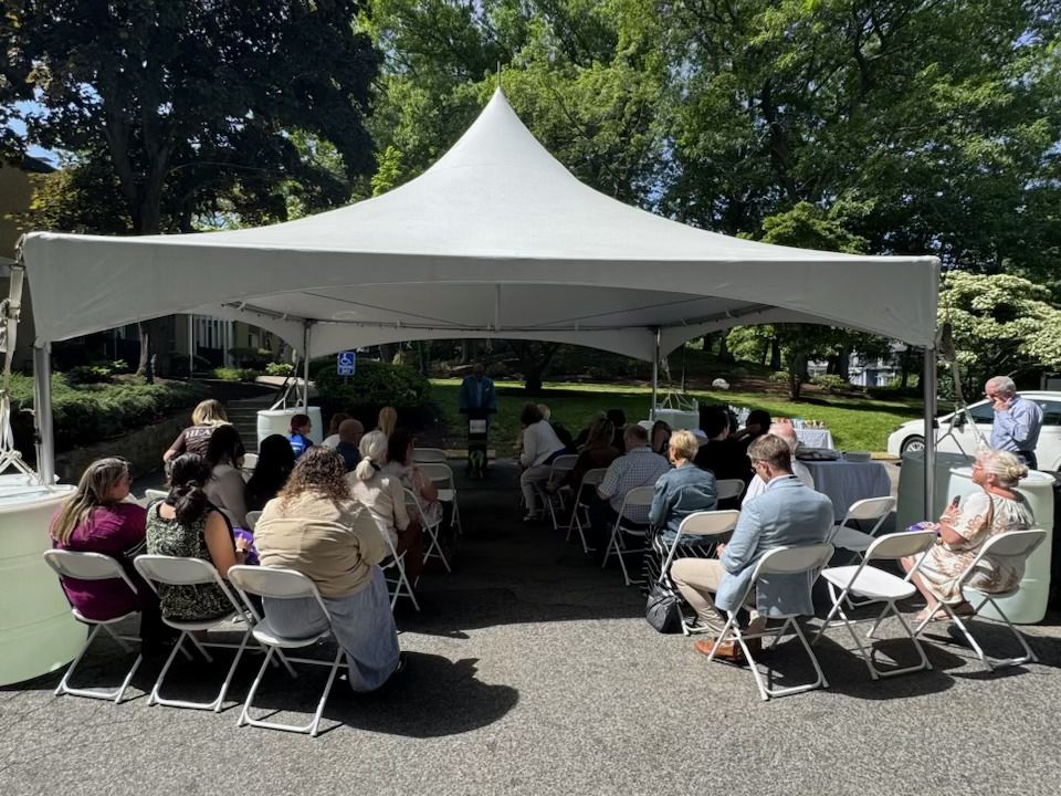 A group of attendees sit under a tent and Tony speaks at the podium.