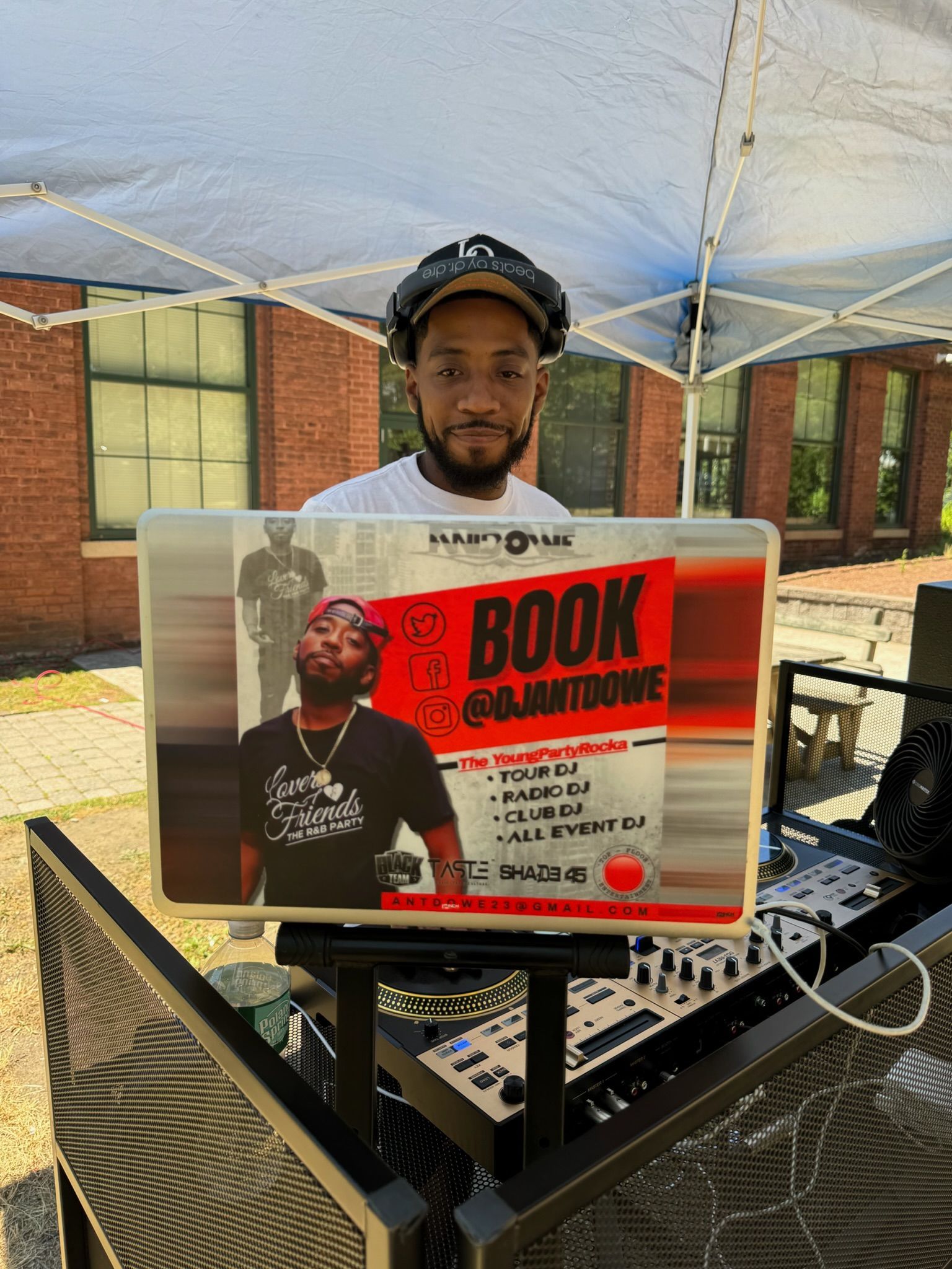 DJ Anthony Dowe standing under a tent with his DJ equipment. There's a sign with his social media username: @djantdowe. He can be found on Twitter, Facebook, and Instagram.
