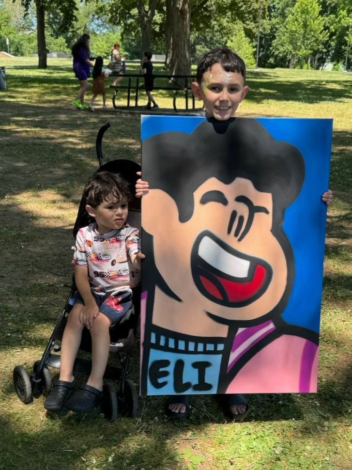 A child stands next to his sibling in a stroller while smiling and holding up his completed graffiti artwork of a cartoon character.