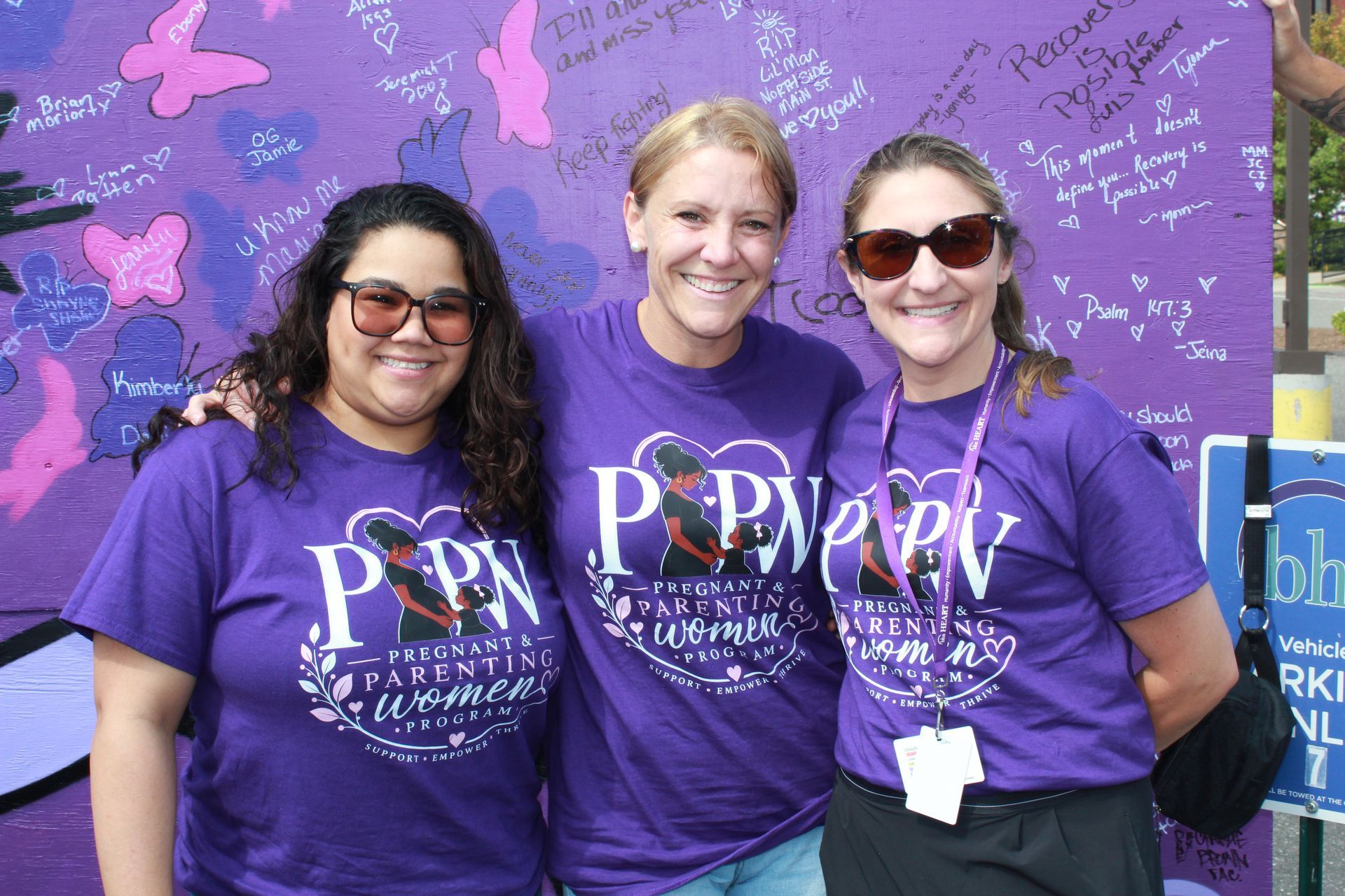 3 women from BHN's PPW program smile for a photo in front of the board for International Overdose Awareness Day while wearing purple shirts from their programs.