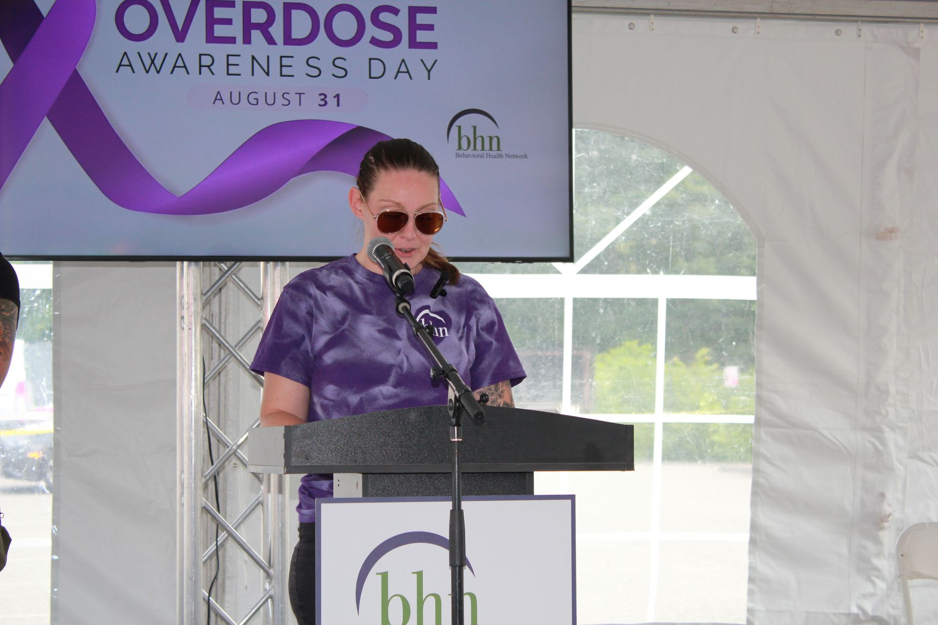 A woman in a purple tie-dye shirt reads a poem at the podium for BHN's International Overdose Awareness Day event.