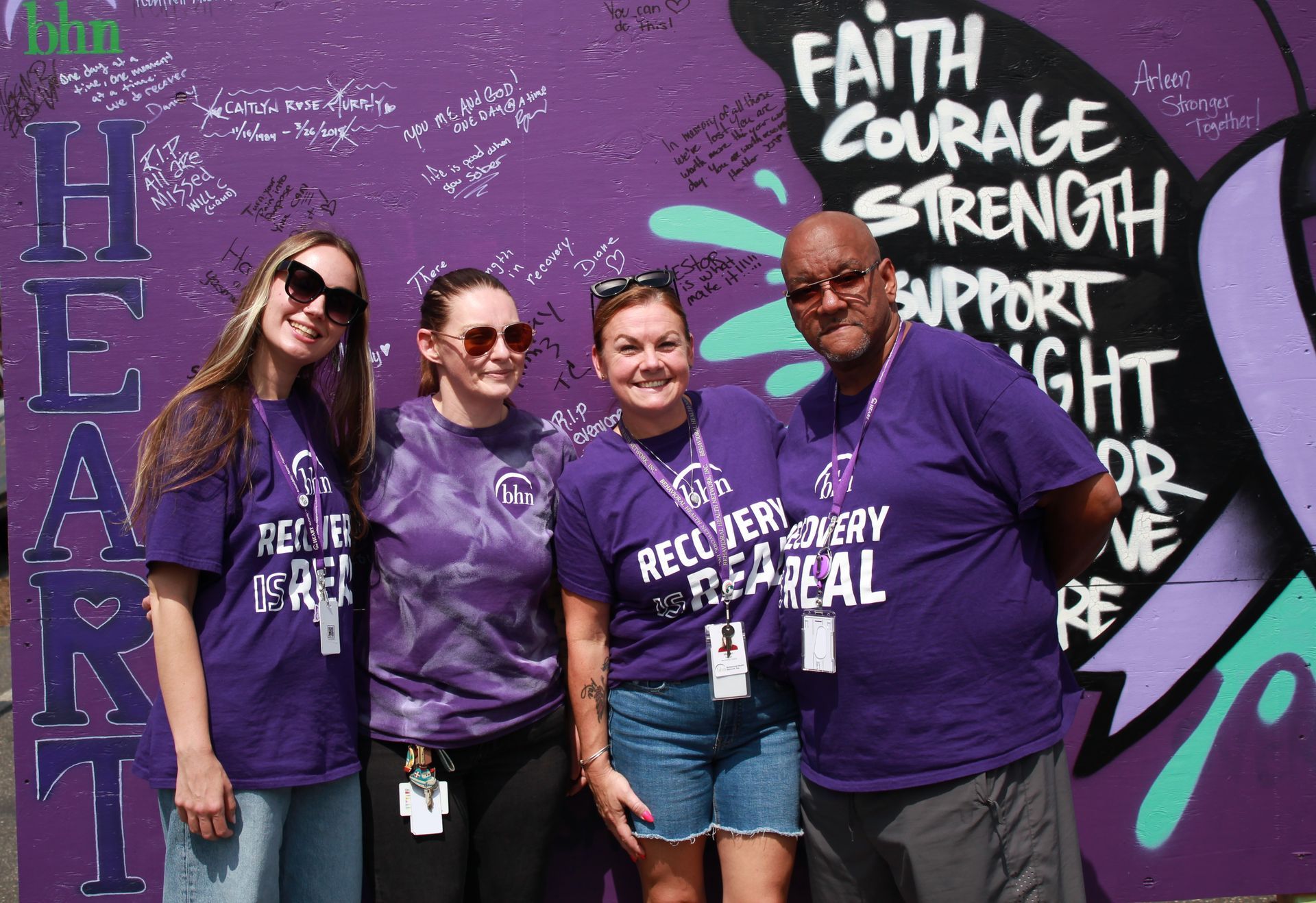 4 of BHN's Recovery Coaches smile and pose for a photo in front of the purple board for International Overdose Awareness Day while wearing purple shirts that say 