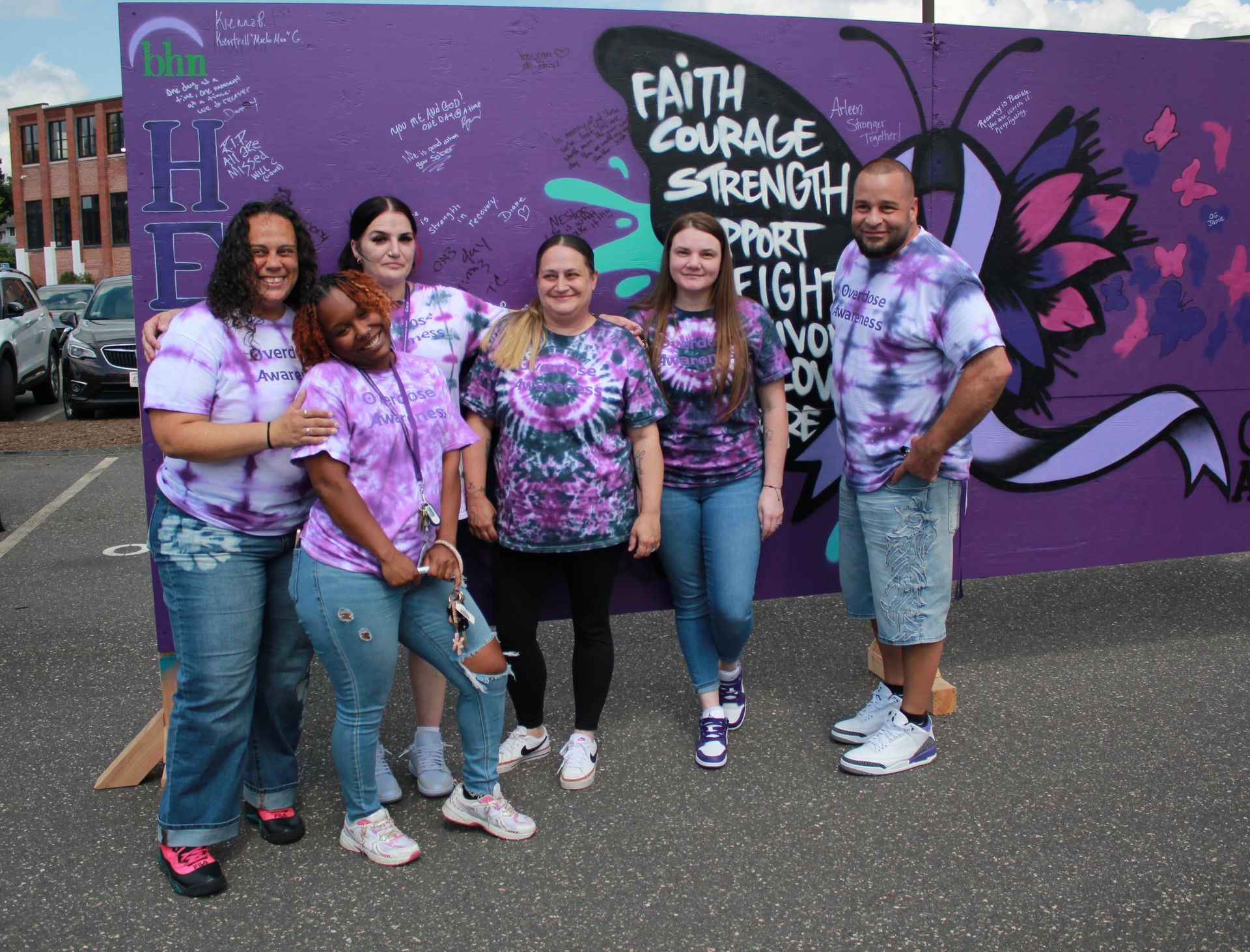 6 of BHN's recovery coaches smile and pose for a photo in front of a purple board that was decorated for International Overdose Awareness Day. They're all wearing purple tie-dye shirts that support overdose awareness. 