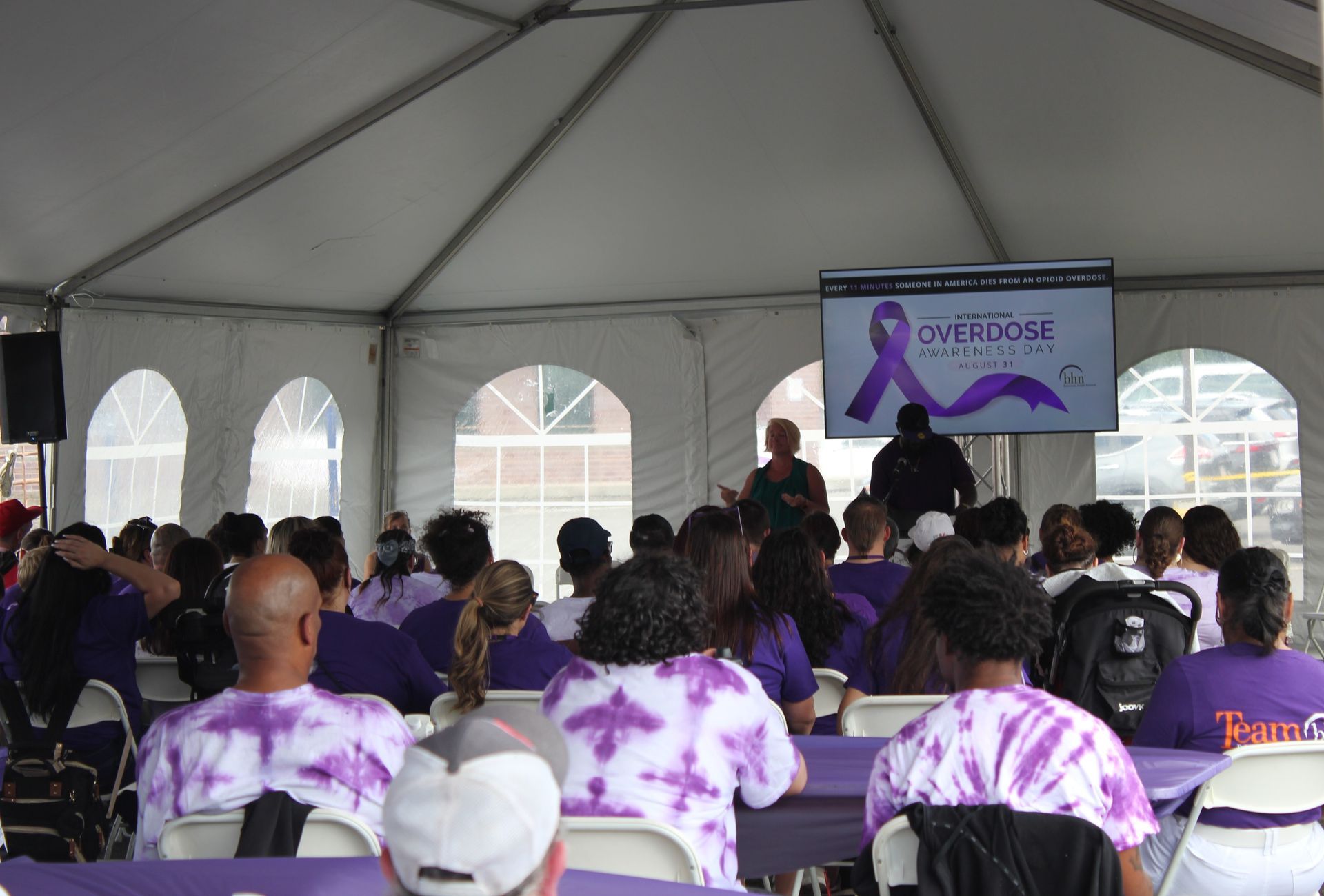 Recovery coaches, BHN staff members, and some recovery clients sit under a tent while listening to speeches for International Overdose Awareness Day.