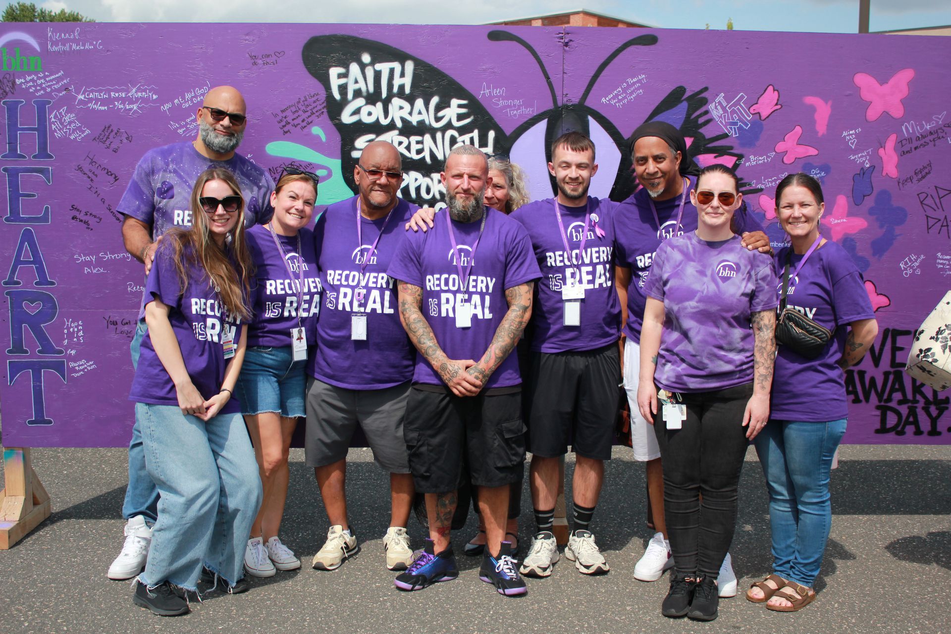 10 Recovery Coaches smile and pose for a photo in front of a large, purple board that was decorated for International Overdose Awareness Day.