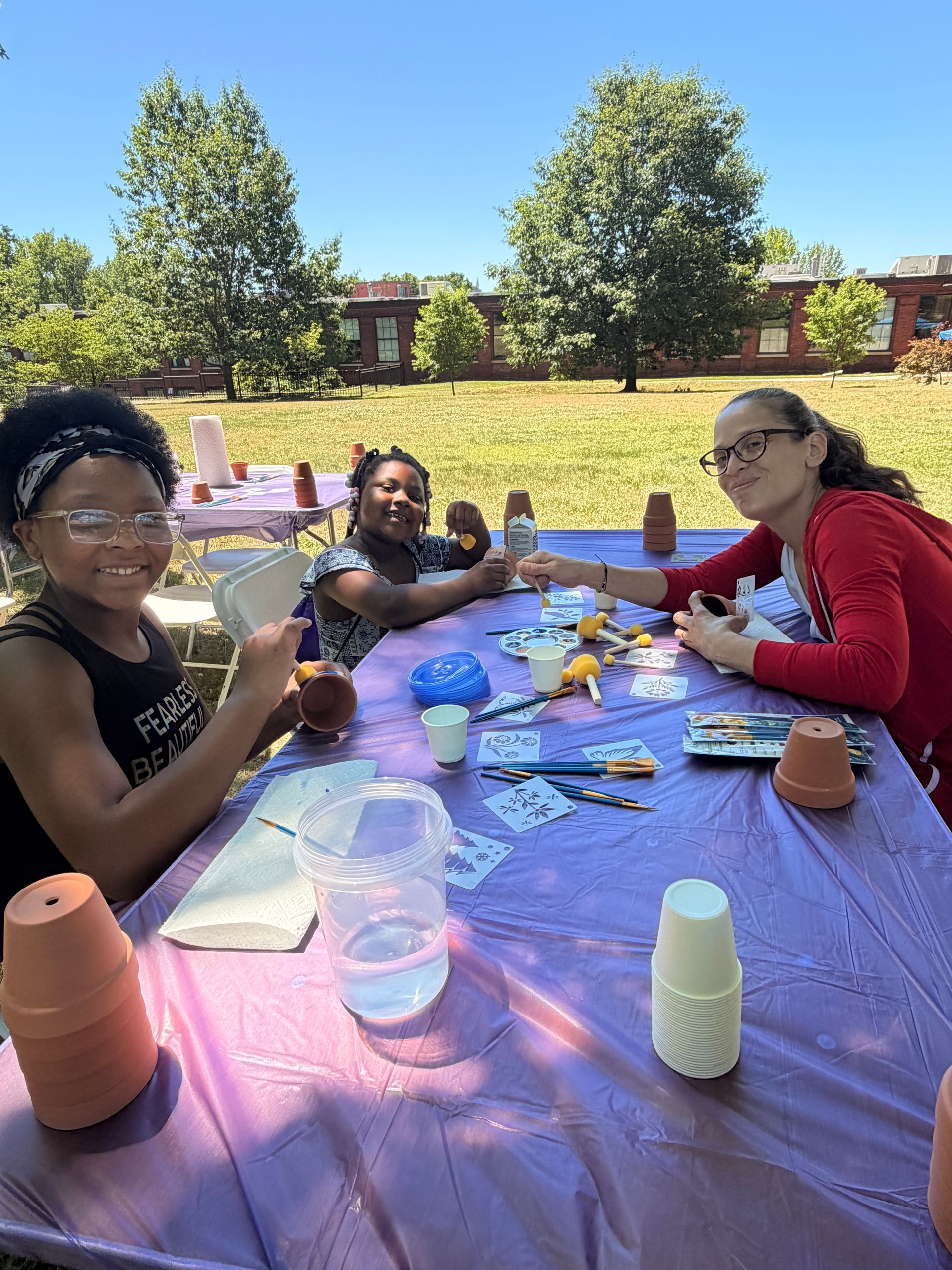 A woman and two children painting flowerpots at a table in the park.
