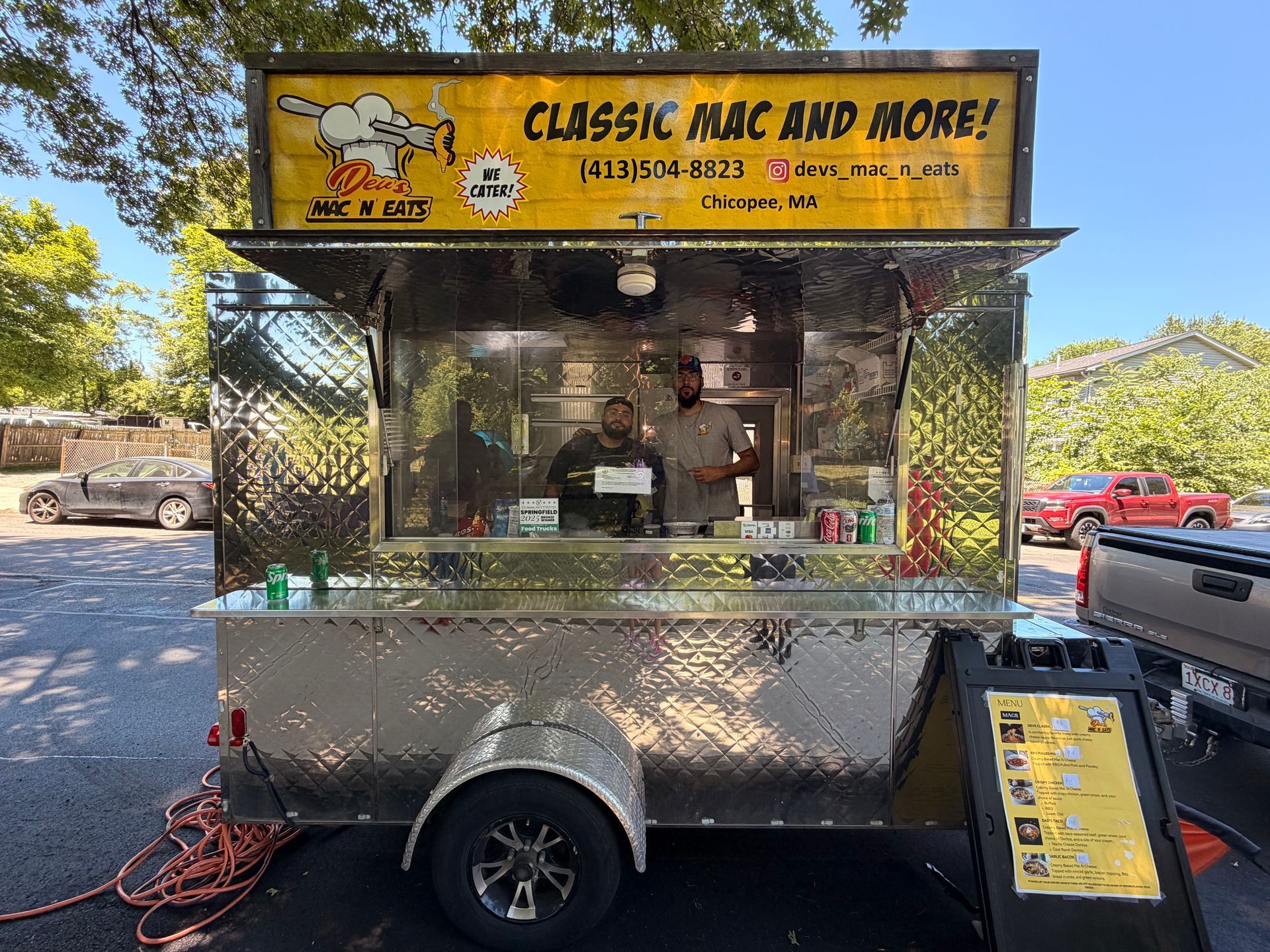 A photo of the owners of the Classic Mac and More food truck and their food truck. Their truck is metallic with a yellow banner at the top that shares their phone number: 413-504-8823 and their Instagram handle: @devs_mac_n_eats