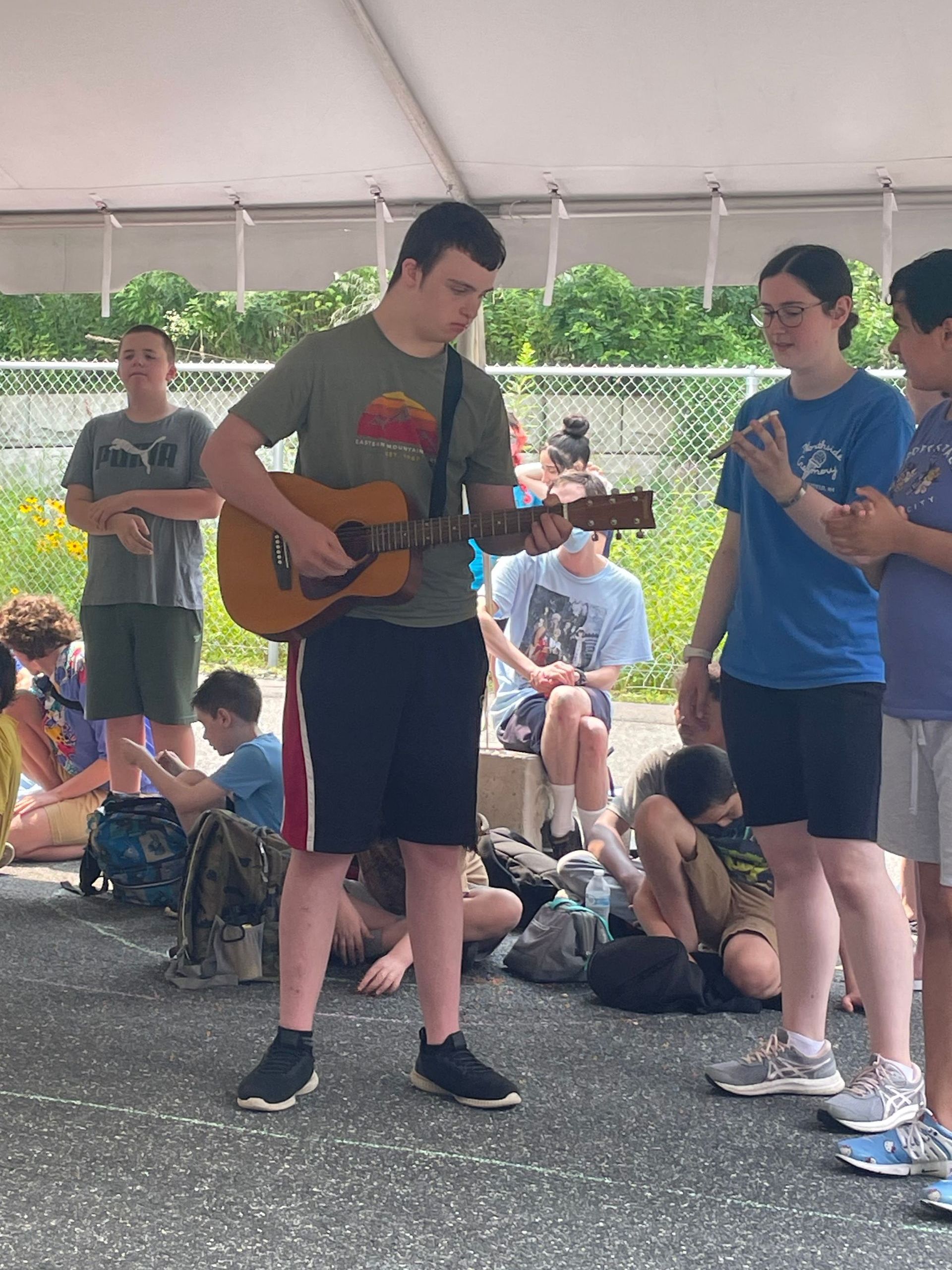Two people play guitar under a tent while others sit and watch behind them