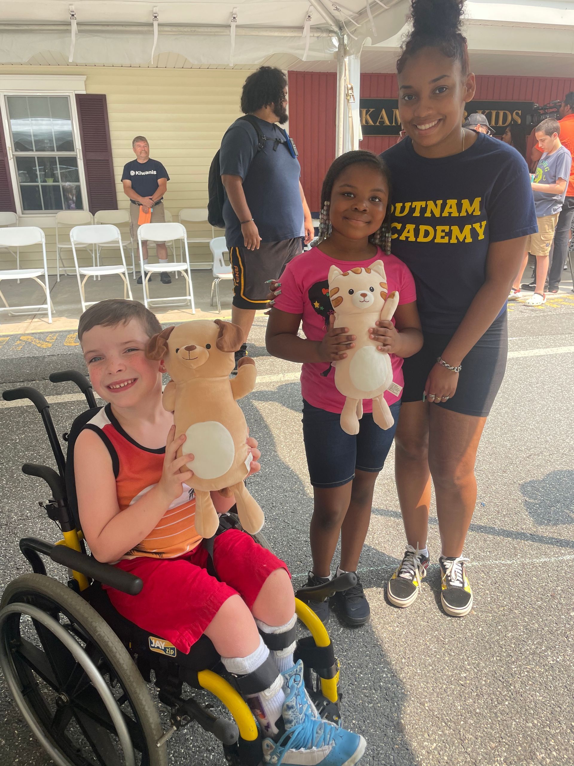 Three children in a covered outdoor area holding stuffed animals, one seated in a wheelchair