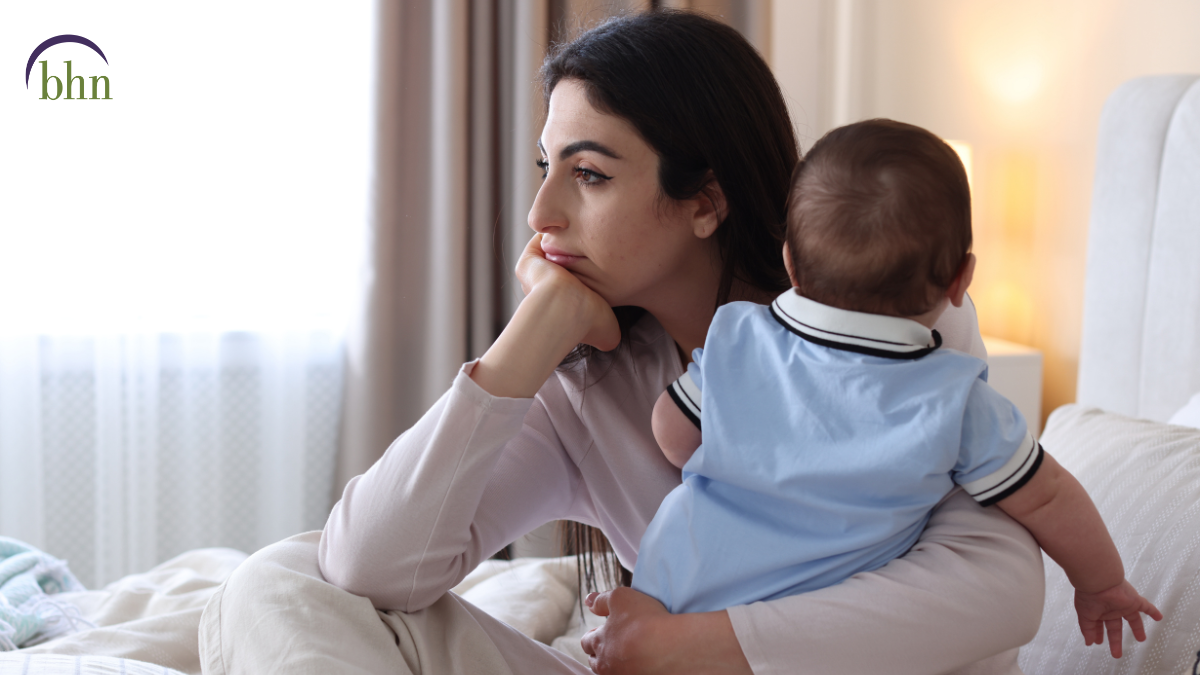 A mother looks off into the distance while sitting in bed with her baby.