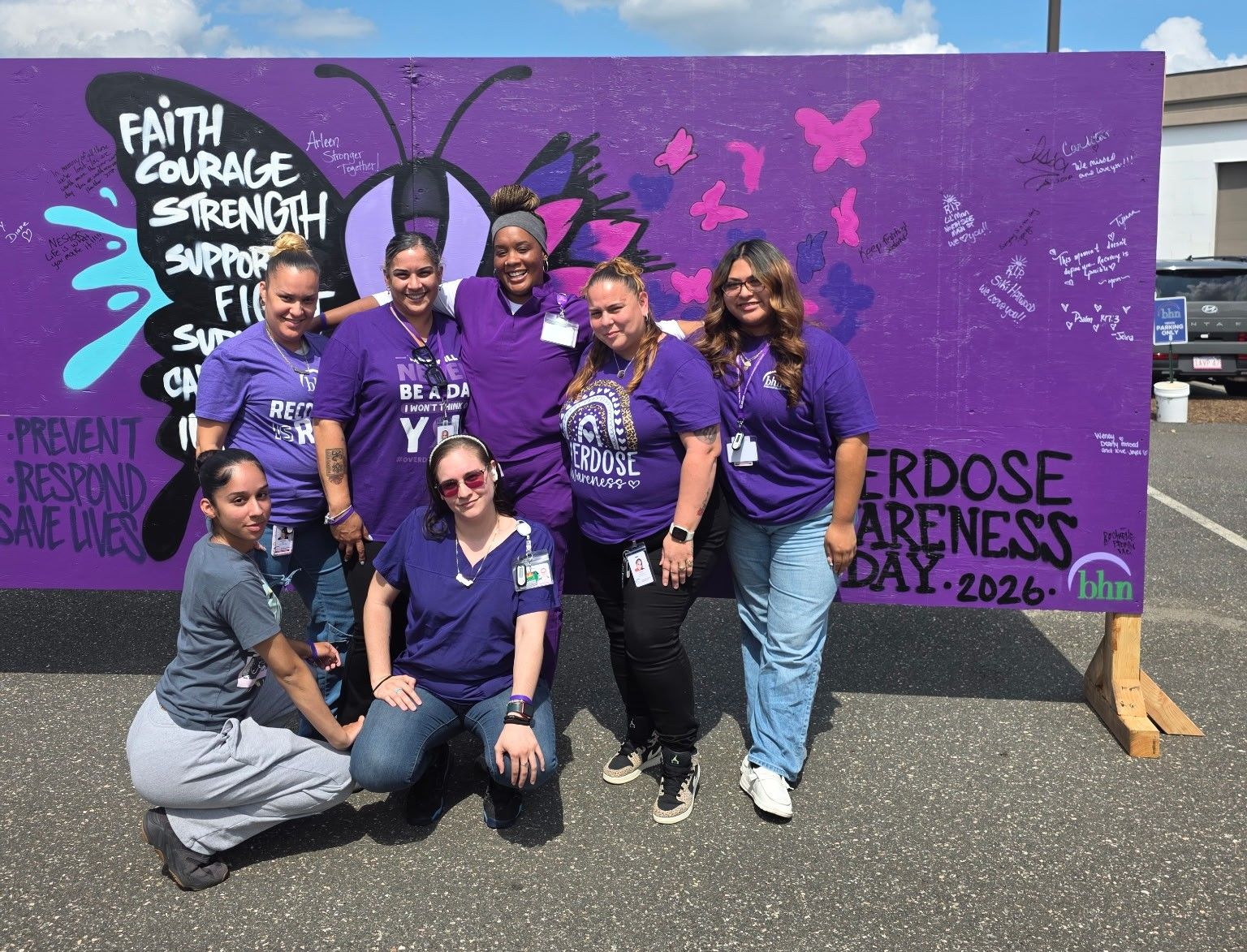 7 Recovery Coaches pose in front of a large, purple board that was decorated for International Overdose Awareness Day.