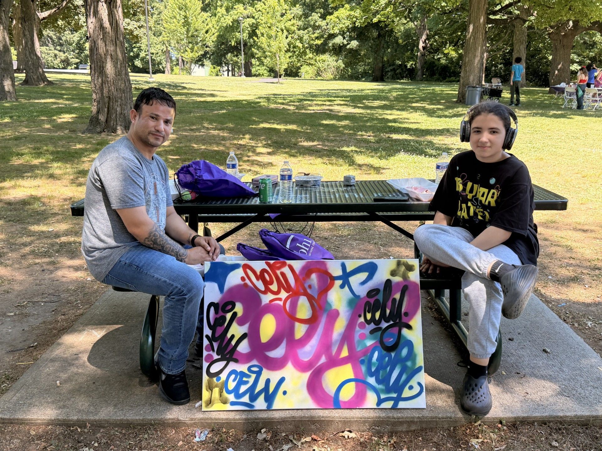 A man and his son sitting at a park picnic table with the colorful graffiti artwork they made displayed in front of them on the ground.