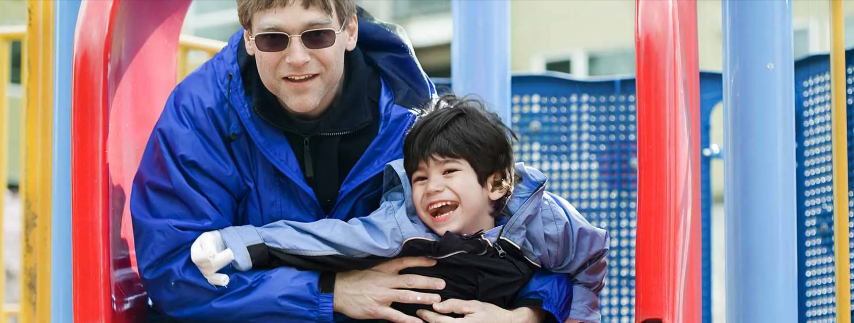 Person in a blue jacket hugging a smiling child at a playground with colorful equipment.