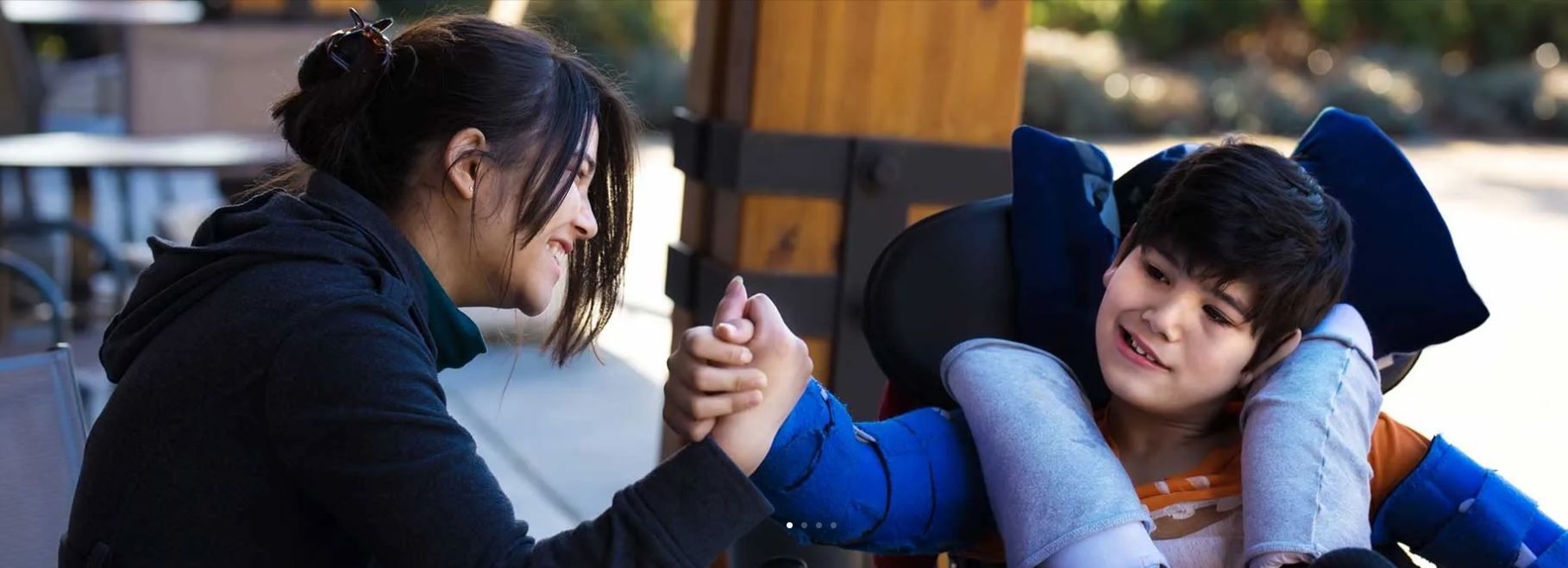 Woman leaning toward a child in a blue jacket, smiling and holding hands indoors.