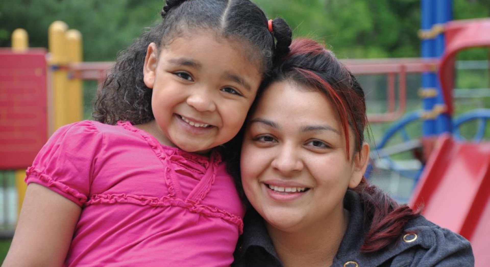 Two people smiling outdoors, one leaning against the other in a colorful playground.