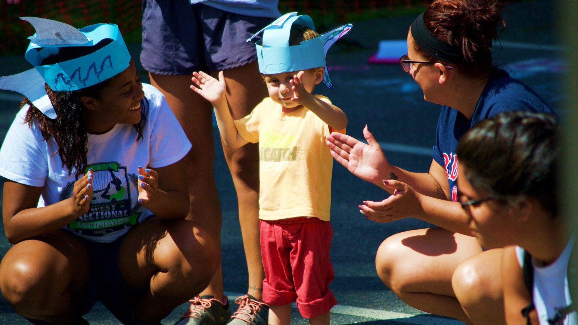 Children wearing paper crowns clap with adults during an outdoor activity in sunlight.
