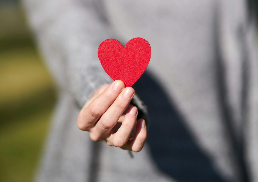 Hand holding a small red felt heart against a blurred gray background