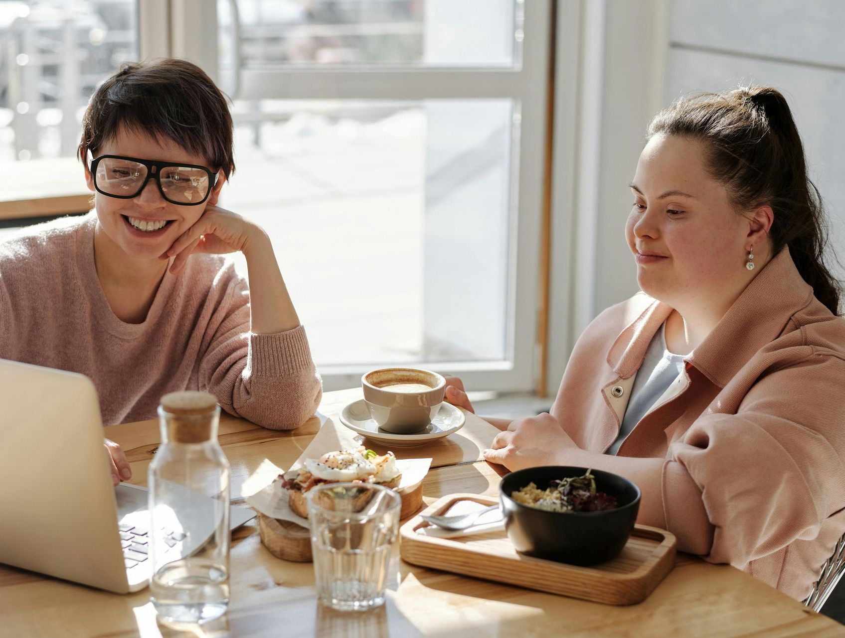 Two people smiling at a laptop during a meal at a sunlit table with tea and bowls