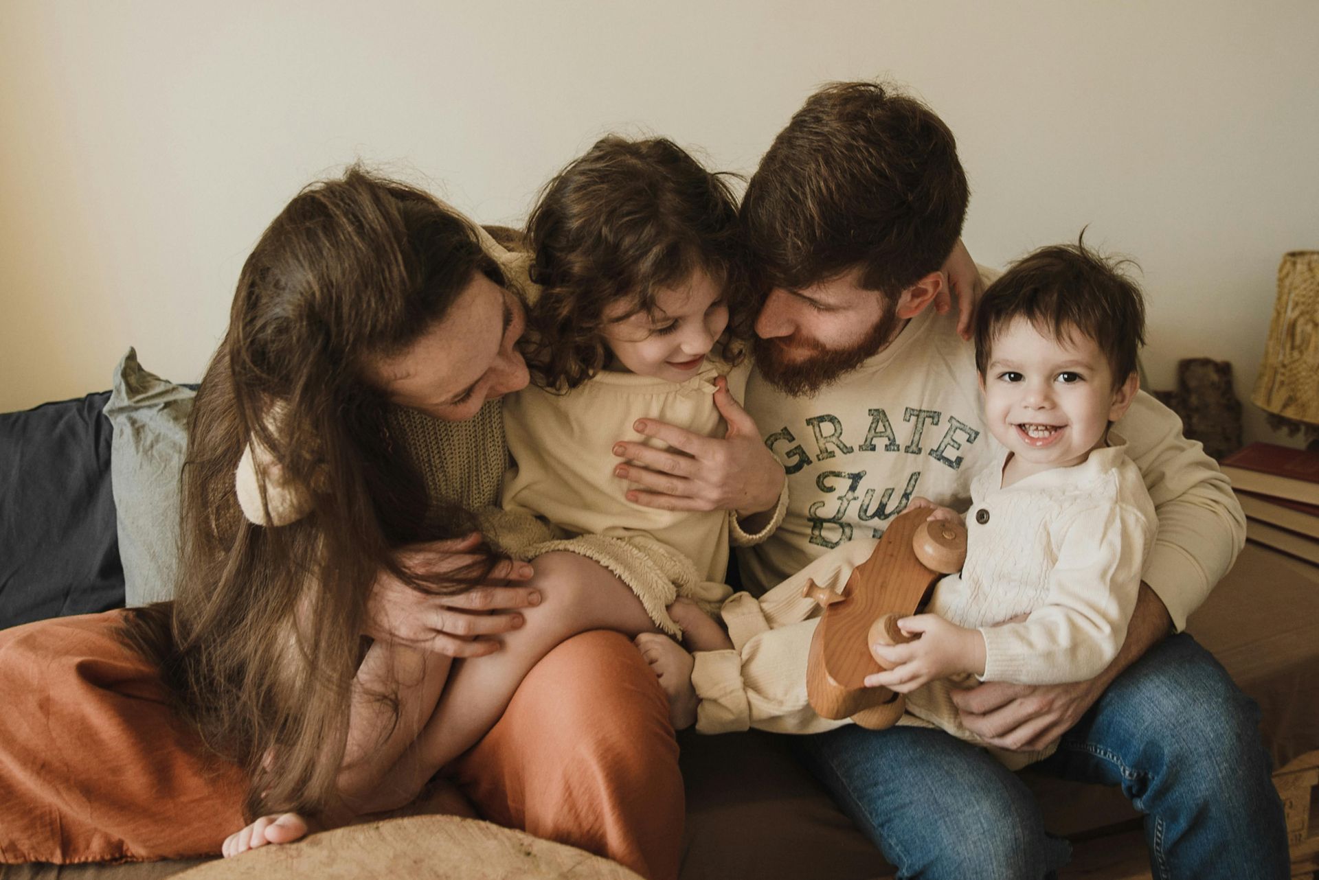 Four people cuddle on a couch, with a toddler in a cream outfit sitting on an adult’s lap.