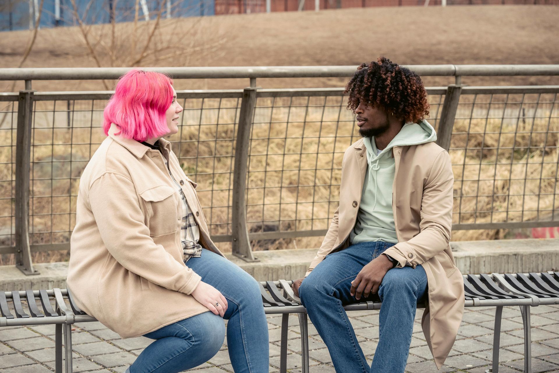 Two people sit facing each other on a bench outdoors, one with bright pink hair, in a muted park setting.