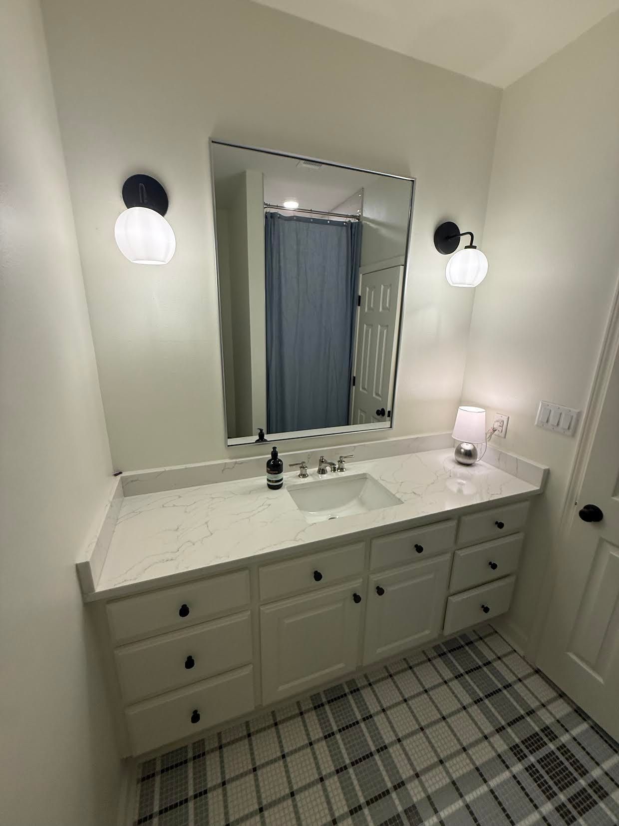 Bright white bathroom with double vanity, wall sconces, mirror, blue shower curtain, and checkered floor