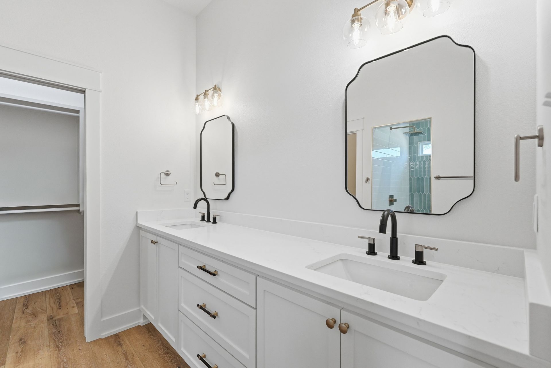 Bright white bathroom vanity with double sinks, black fixtures, framed mirrors, and a walk-in closet visible beside it