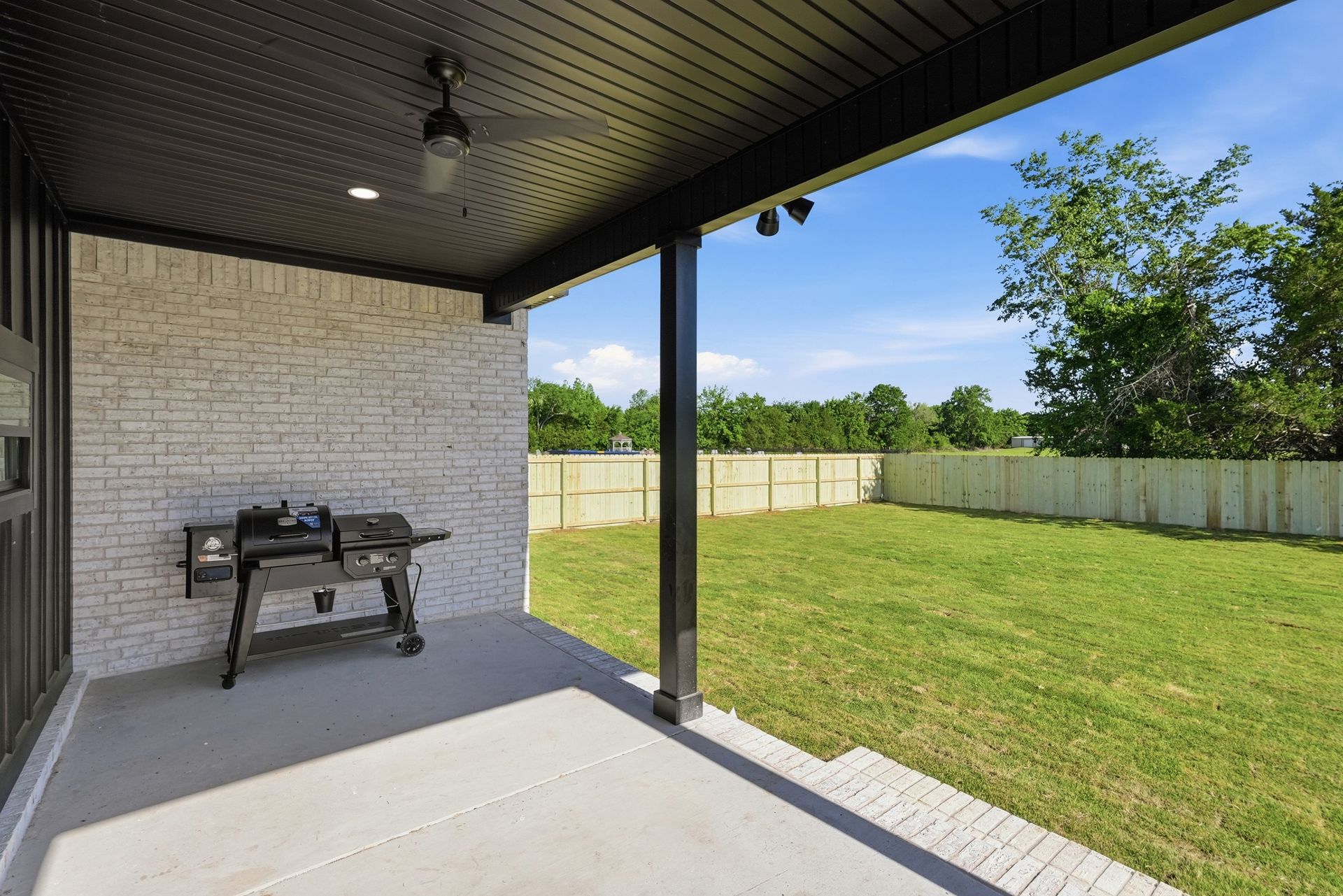 Covered patio with a grill overlooking a grassy backyard and white fence