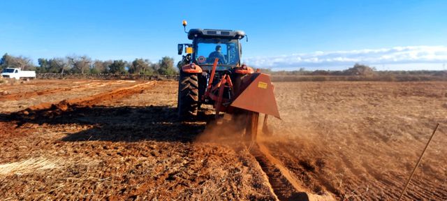 Trattore blu che ripulisce un campo di terra arida con una lama anteriore, sollevando polvere.