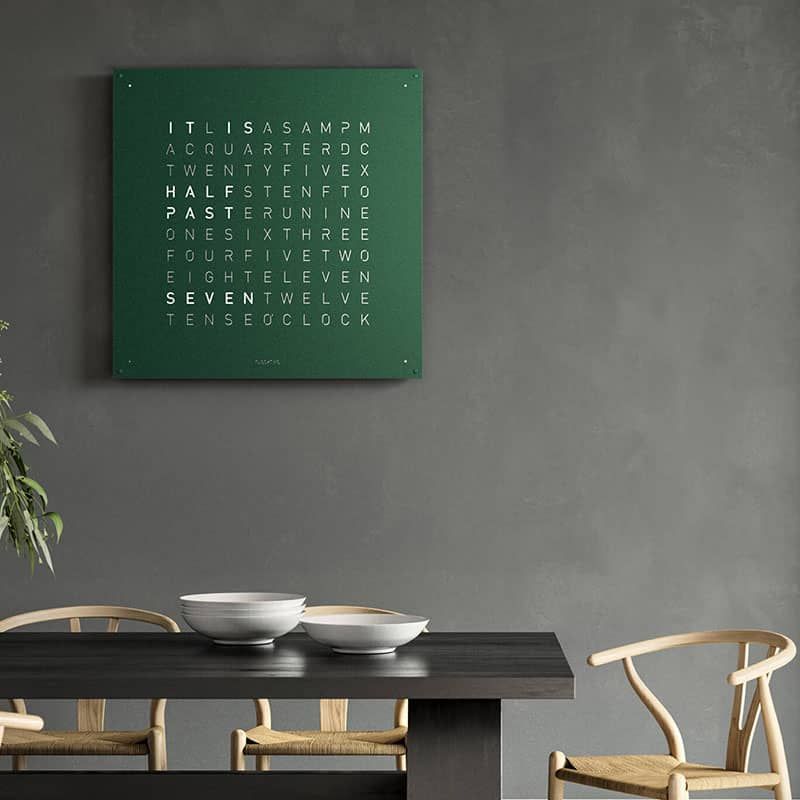 Groene klok met tekst op een grijze muur boven een zwarte tafel met schalen en stoelen.