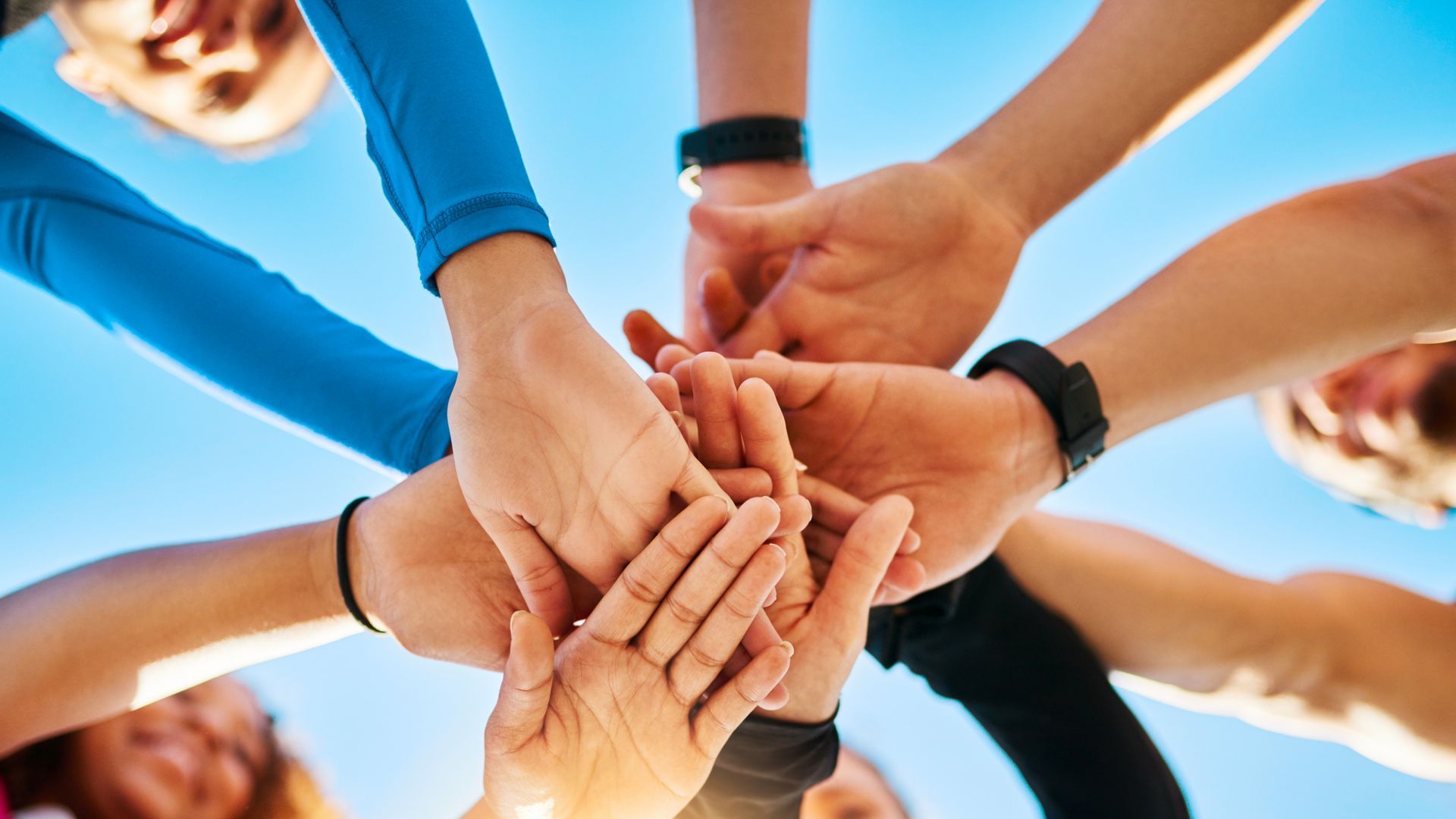 Hands of a diverse group stacked together against a blue sky, showing teamwork and unity