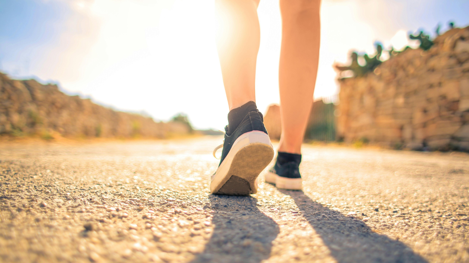 Low-angle view of a person walking on a sunlit dirt path, with sneakers and long shadows