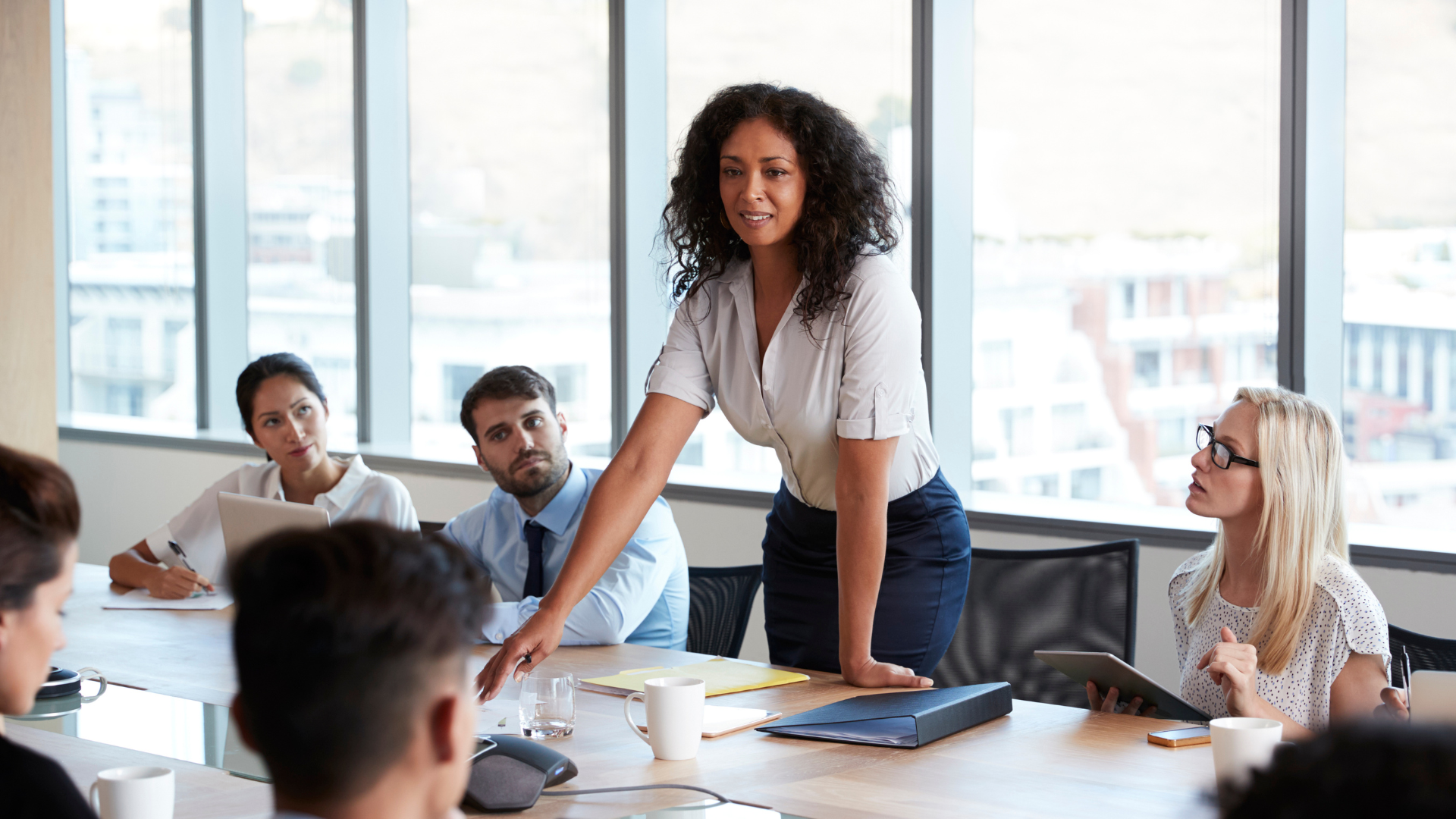 Woman standing at a conference table leading a meeting in a bright office with colleagues seated around her