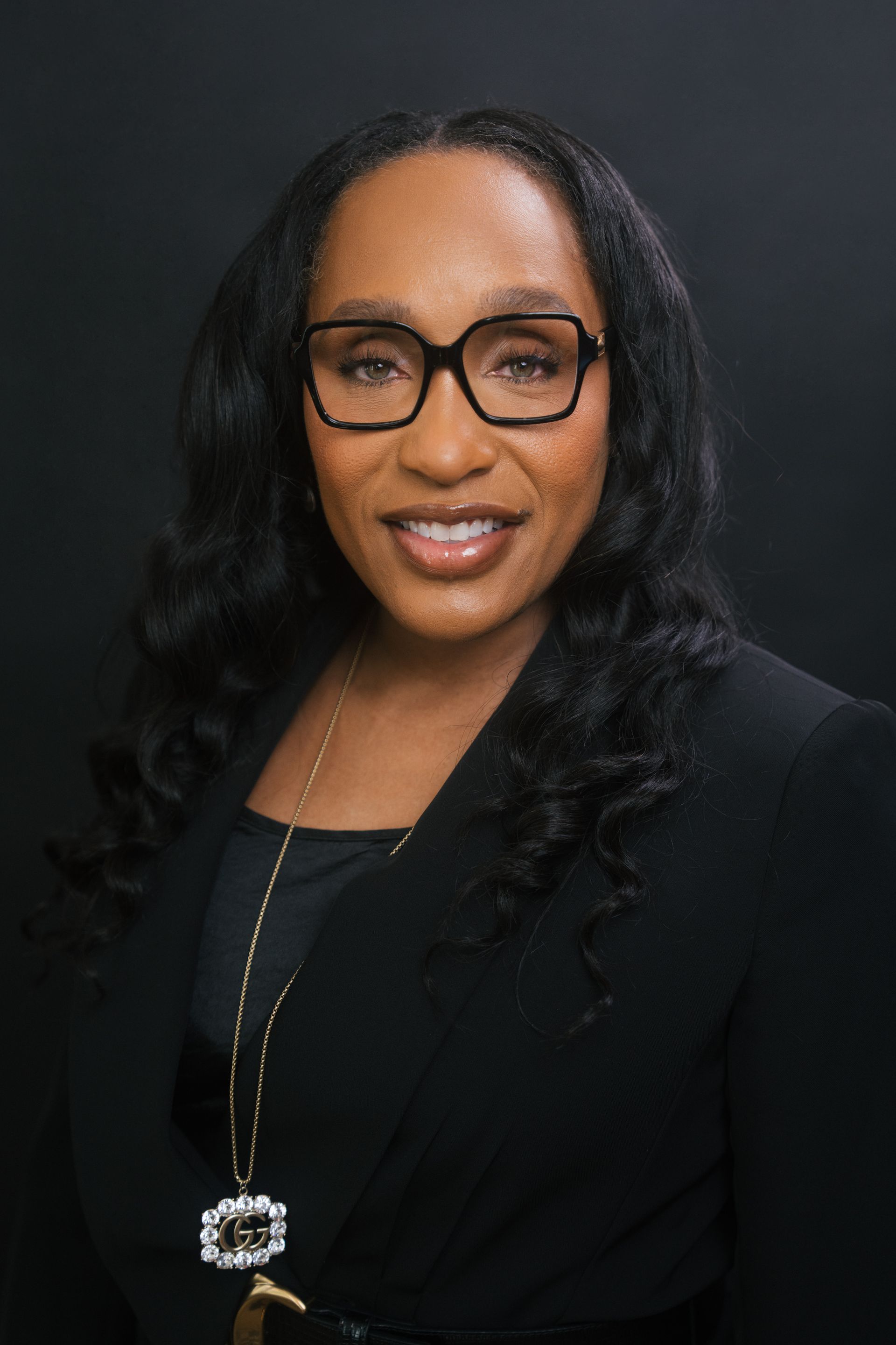 Portrait of a smiling person in a black blazer and glasses against a dark background.