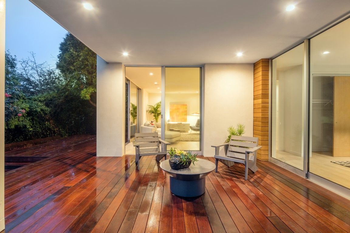 Modern covered patio with wooden deck, lounge chairs, and a central table beside large glass doors.