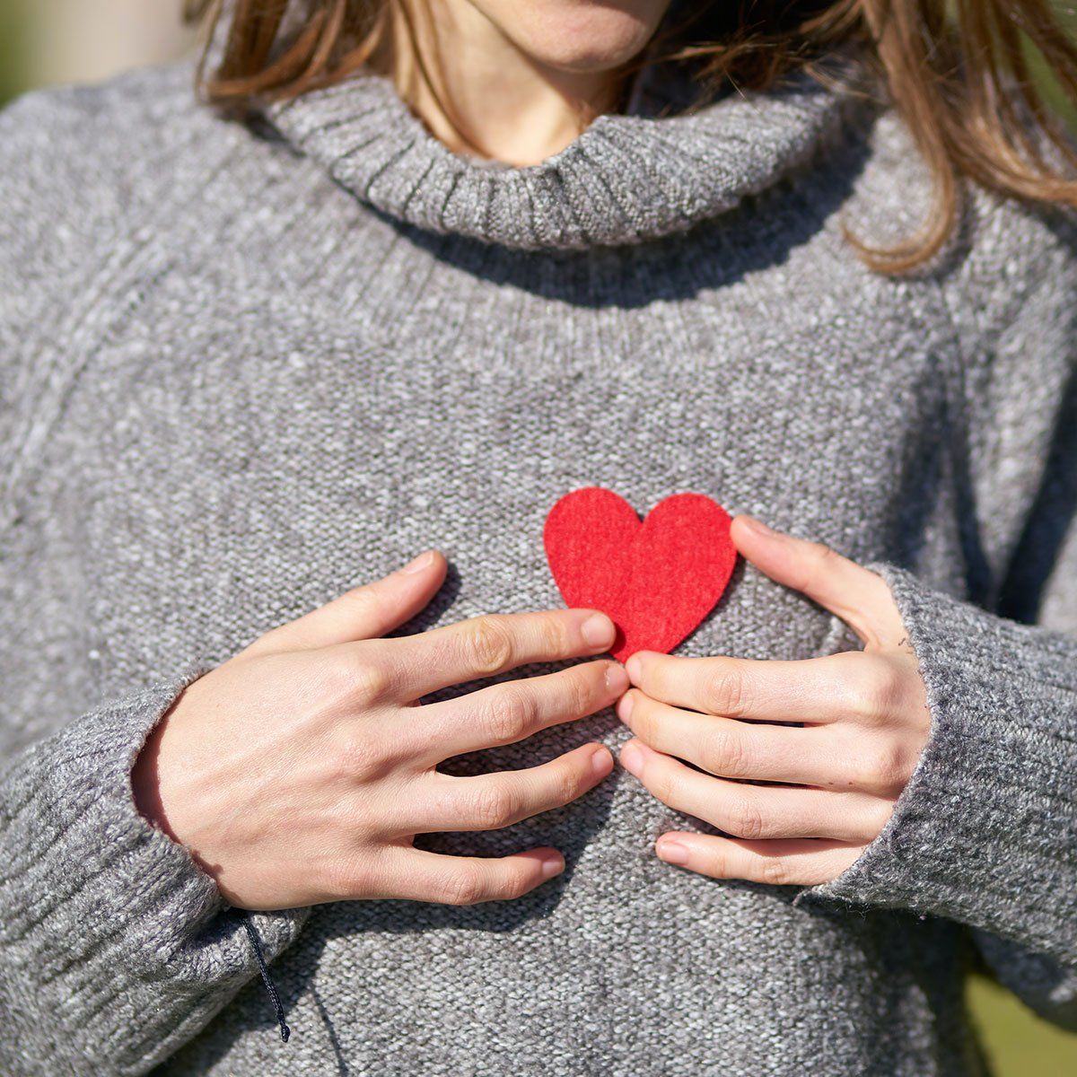 A woman in a grey sweater is holding a red heart in her hands.