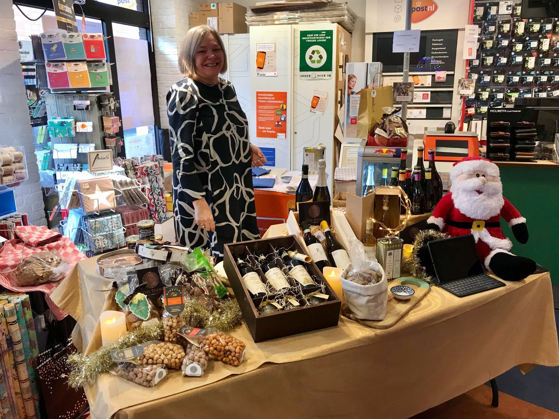 Een vrouw staat achter een tafel met cadeaus, waaronder wijn, in een winkel. Een knuffelkerstman ligt er vlakbij.