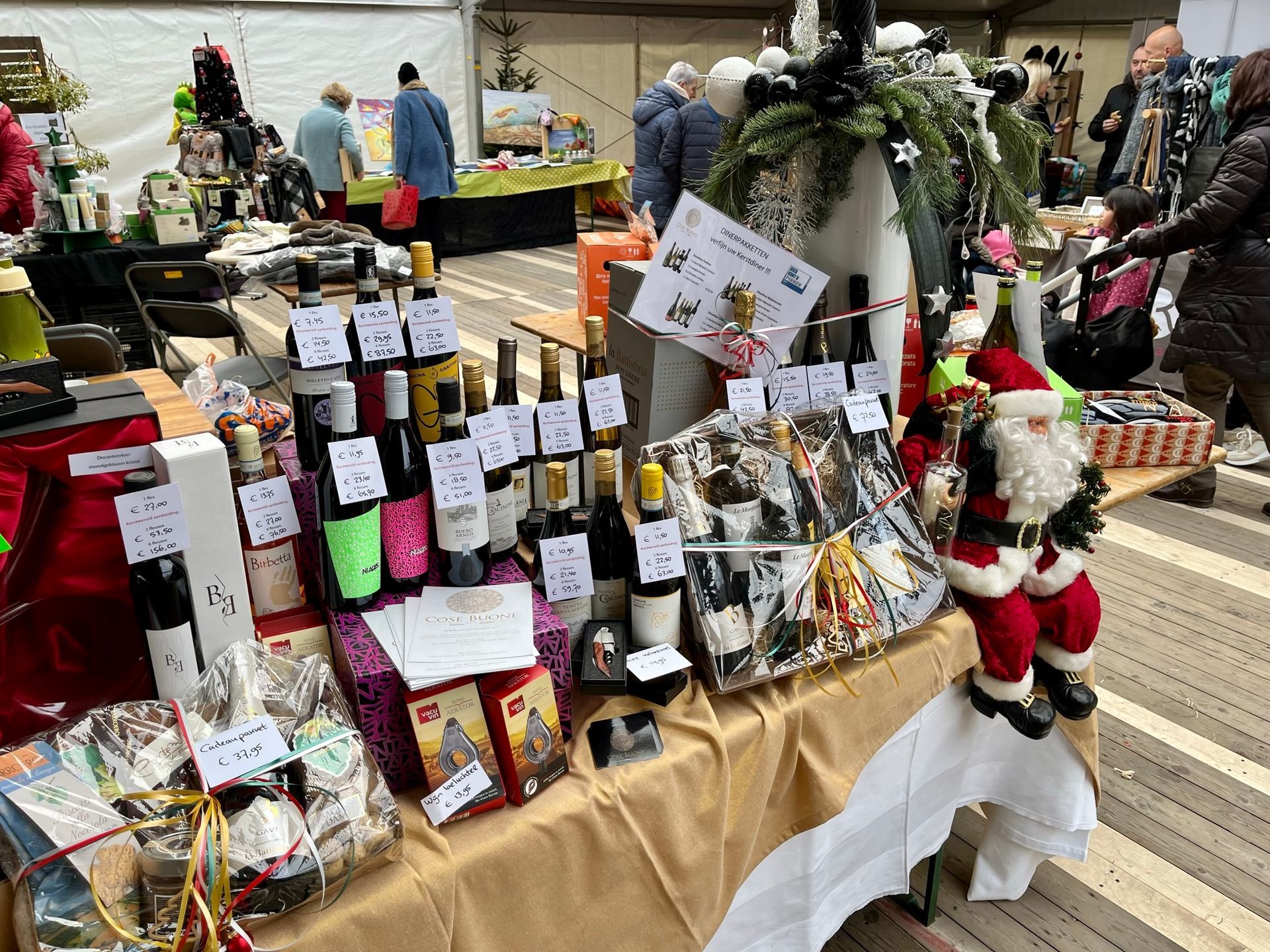 Marktkraam met wijnflessen, cadeaumanden en kerstmanversiering; mensen kijken rond.