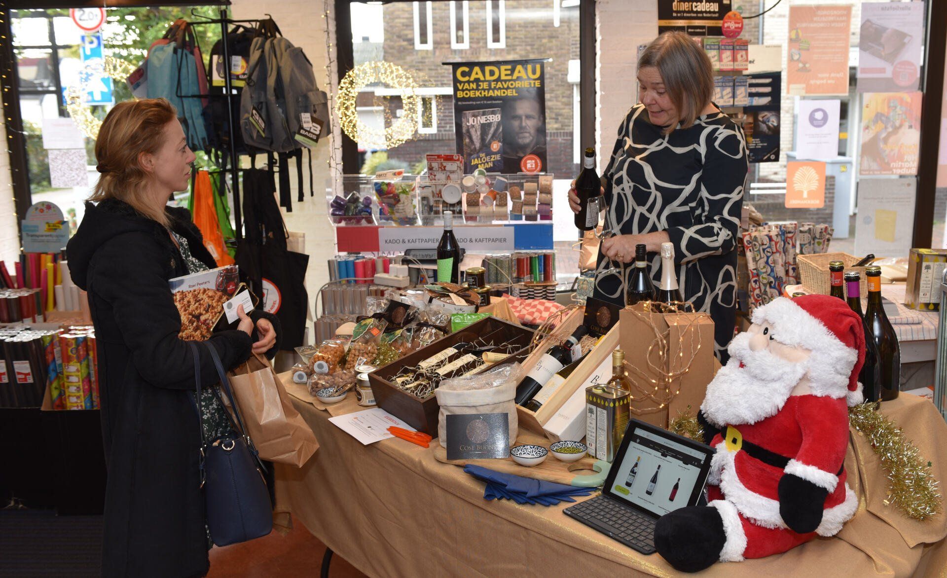 Een vrouw winkelt bij een kerstmarktstalletje met diverse cadeaus, snacks en een kerstmandecoratie.
