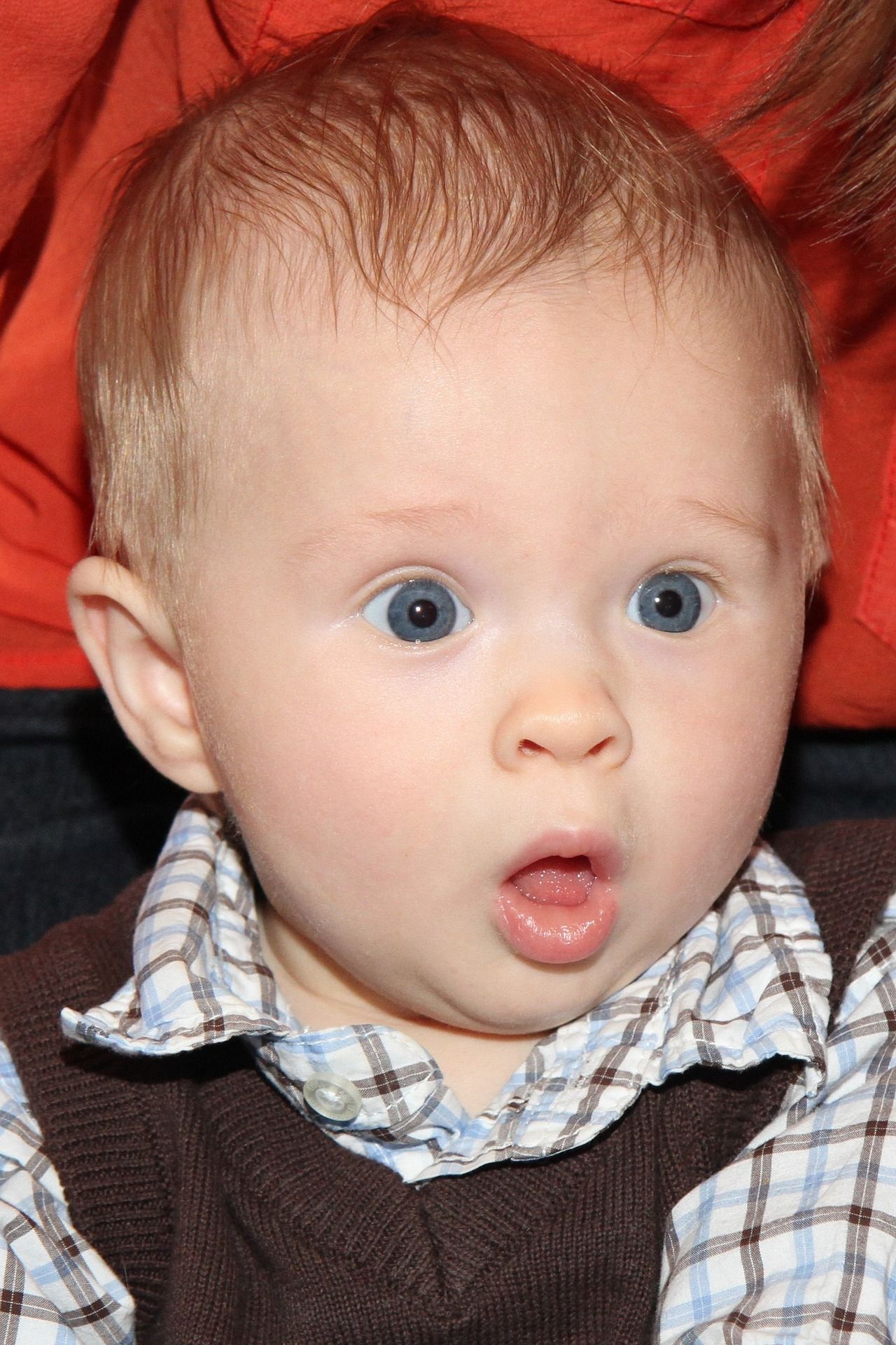 Baby met licht haar en blauwe ogen, een verraste uitdrukking, open mond, gekleed in een bruin vestje en een geruit shirt.