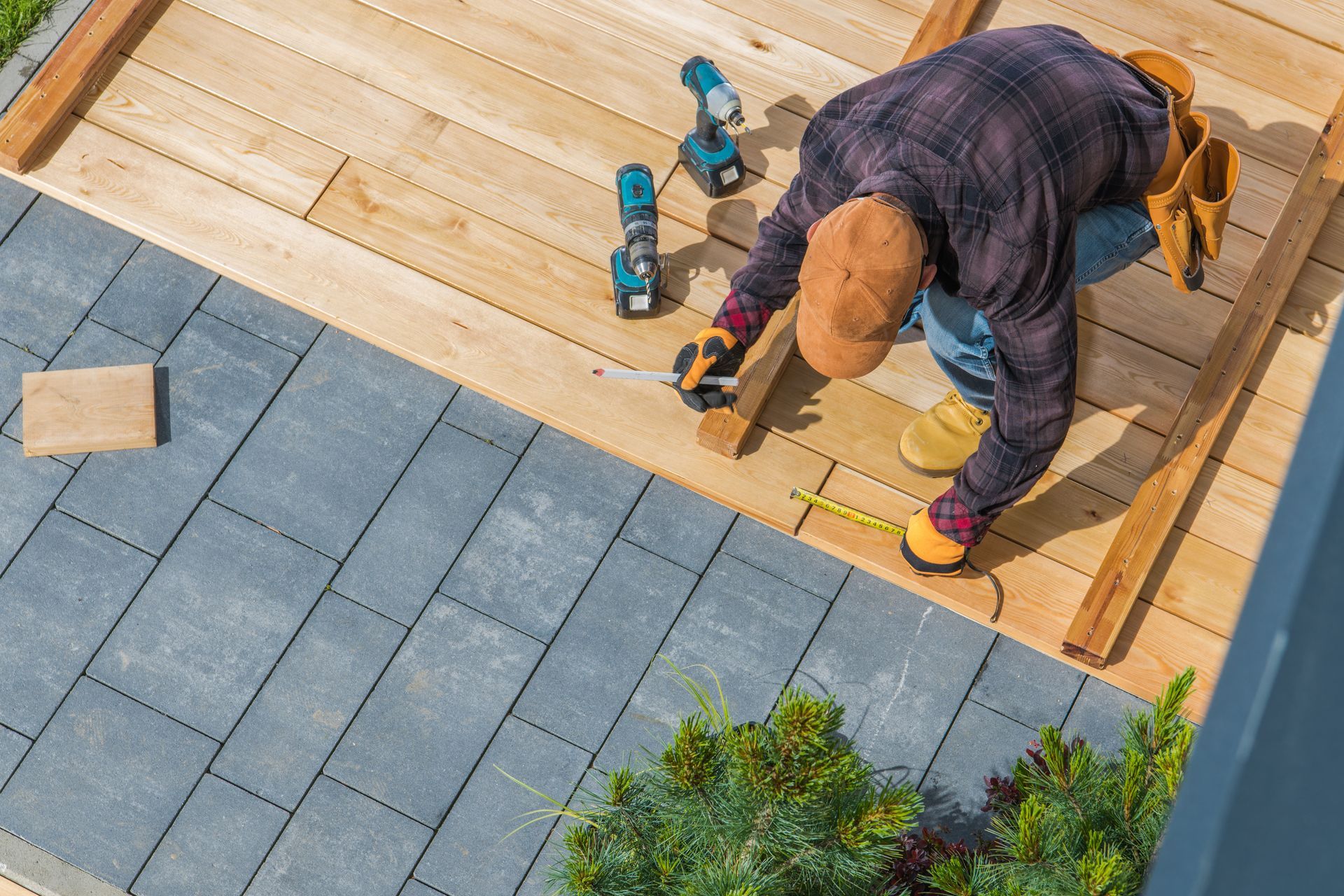 Worker laying wooden deck boards beside gray pavers, using a drill outdoors.