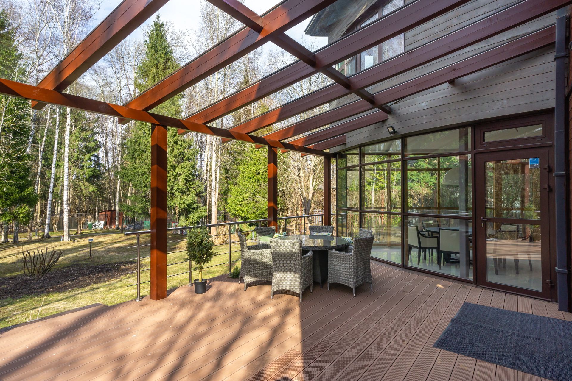 Covered outdoor patio with dining table, chairs, and glass walls beside a wooded yard