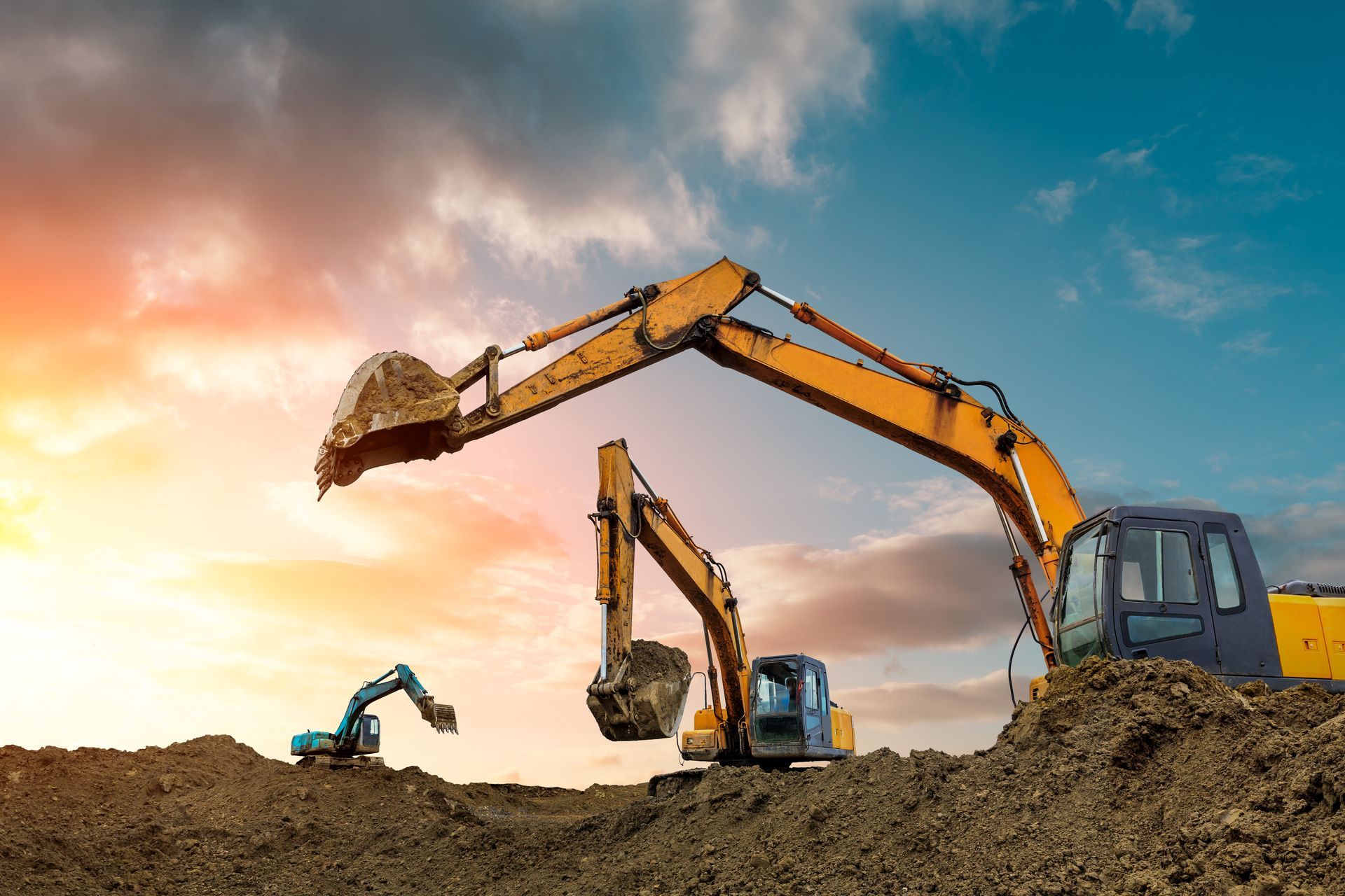 Construction excavators working on a dirt mound at sunset under a colorful sky